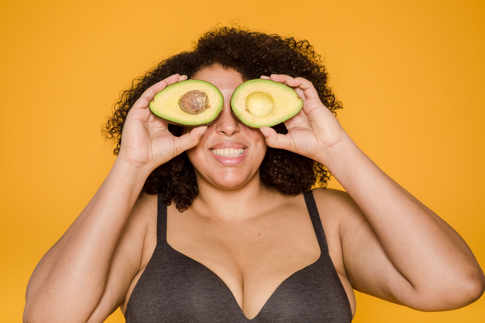 Plump African American woman in bra with curly hair and toothy smile covering eyes with avocado halves