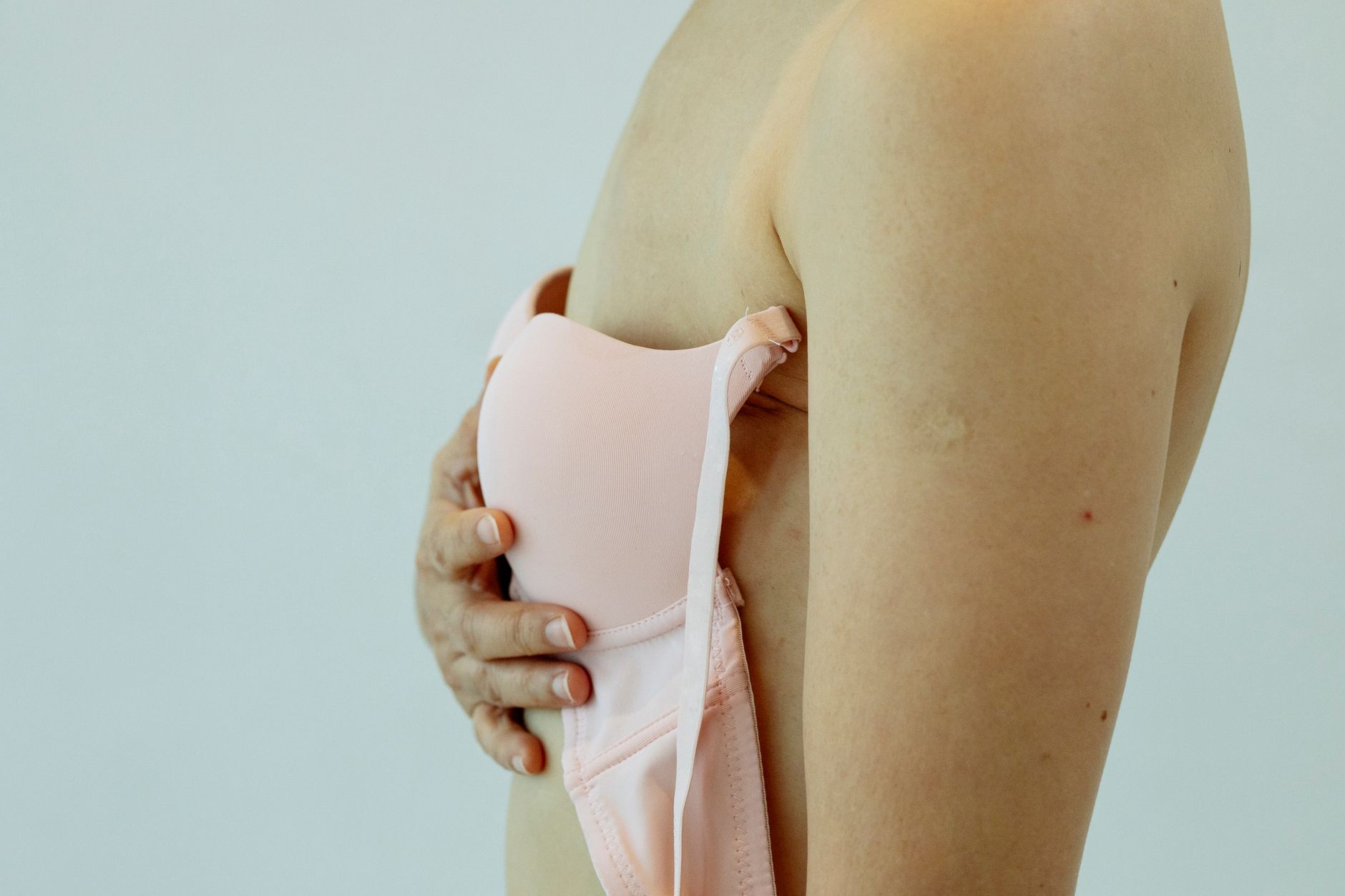 Side view of crop unrecognizable woman with cancer of breast holding pink bra and standing in studio with light walls