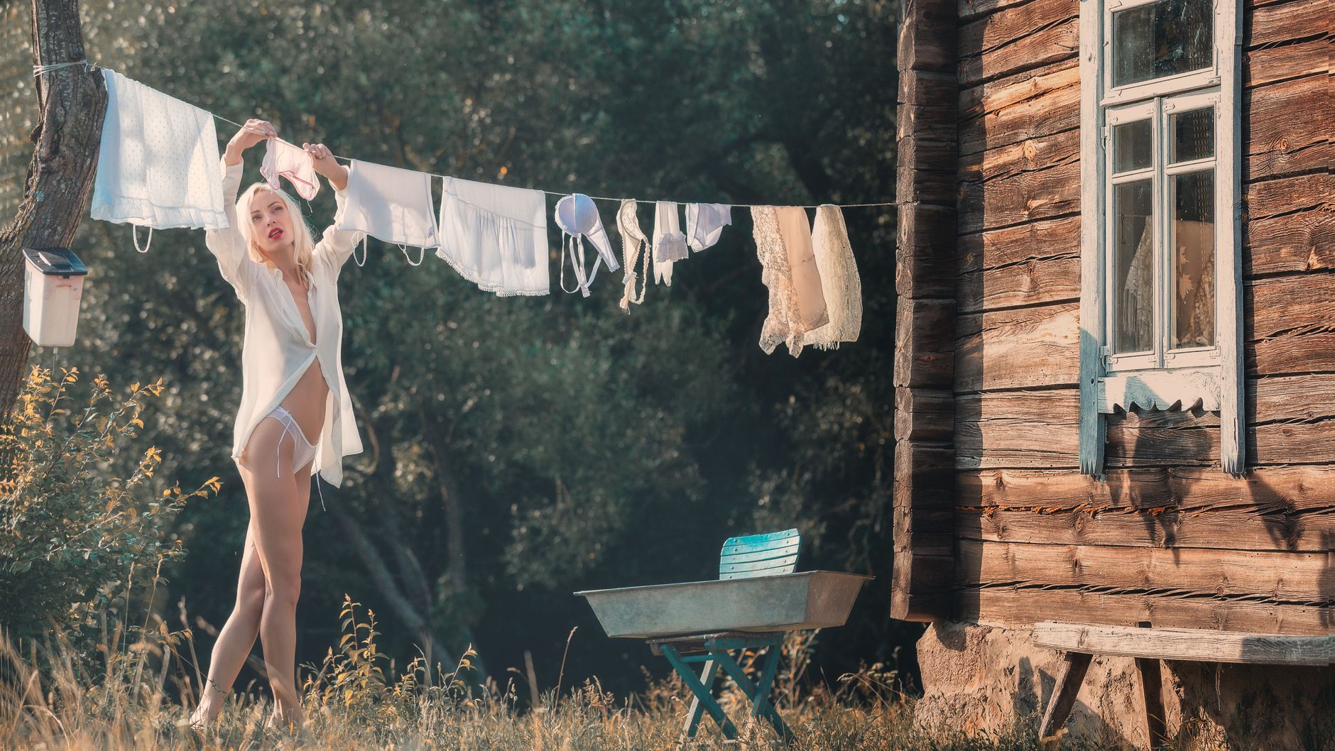 Alluring young female in panties and translucent shirt hanging washed clothes on line while spending summer holidays in countryside