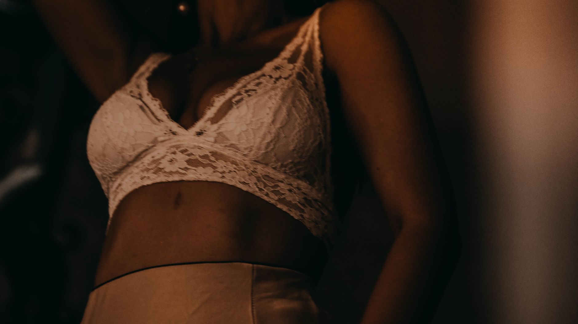 Crop unrecognizable fit young female in sexy lace bra standing near wall in dark room