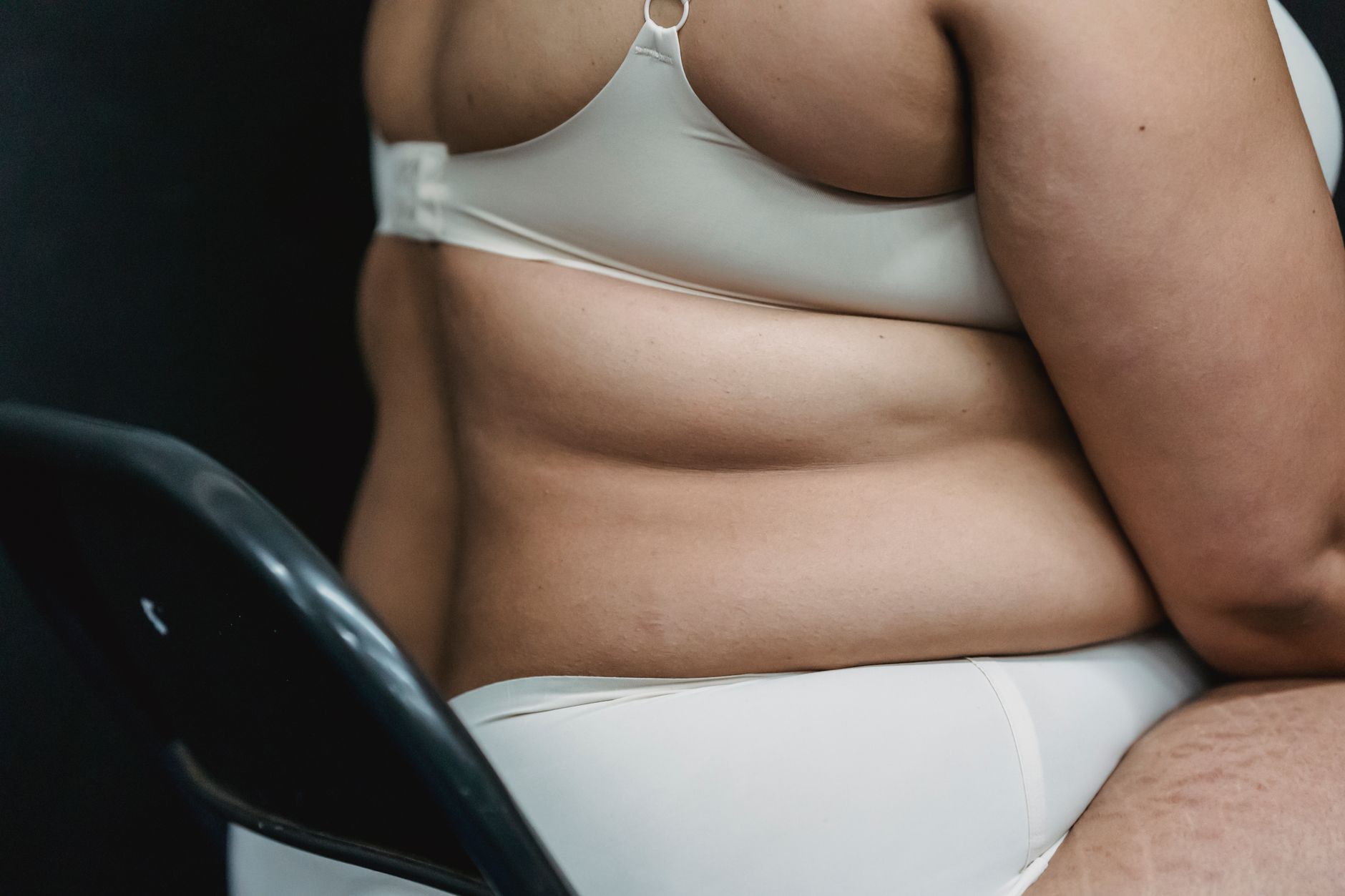 Crop faceless plump woman in white underwear sitting on chair in light studio on dark background