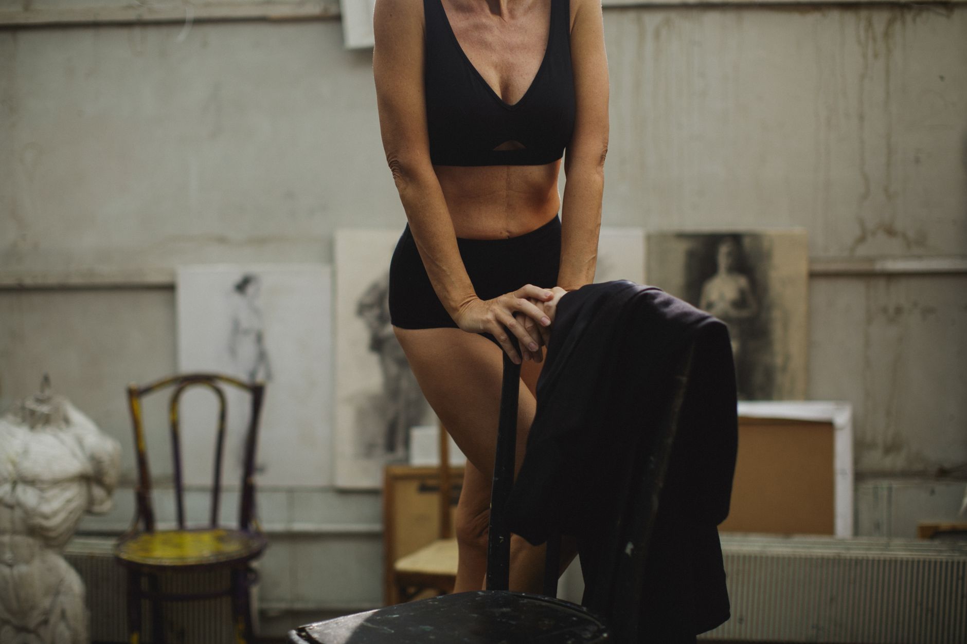 Female model in studio, wearing black underwear with artistic sketches and sculptures in the background.