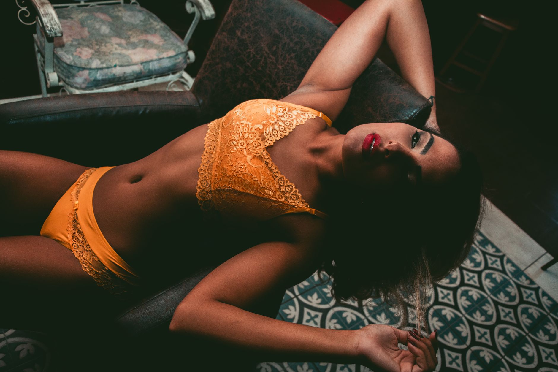 A beautiful woman in yellow lace lingerie lounges elegantly on a patterned armchair indoors.