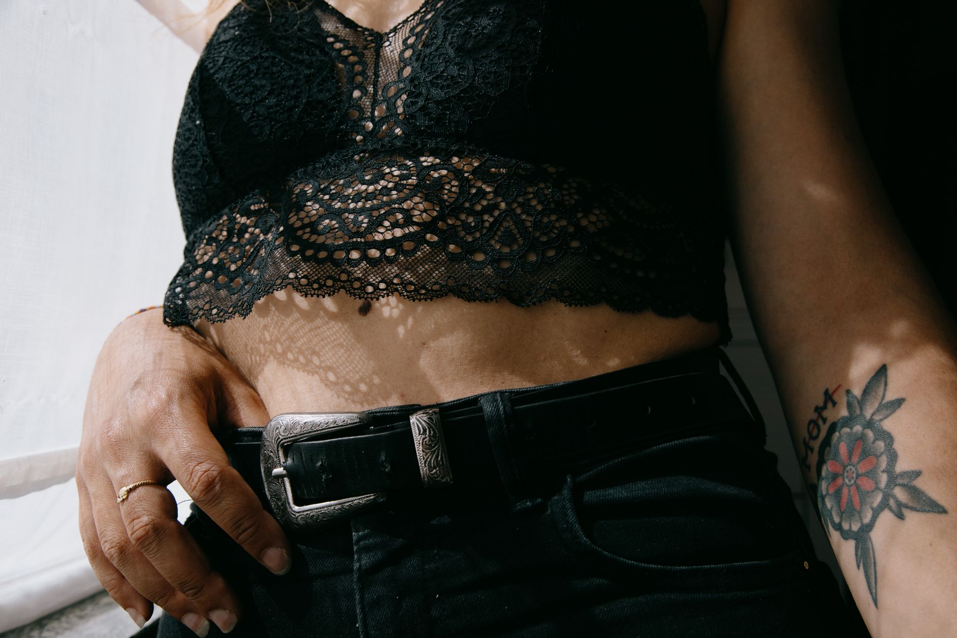 Crop faceless female with tattoo and ring in lace bra and black jeans standing under sunlight