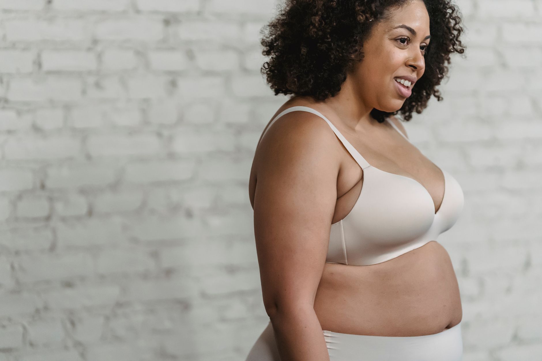 Side view of crop plump African American female with curly hair wearing lingerie standing near white brick wall in studio