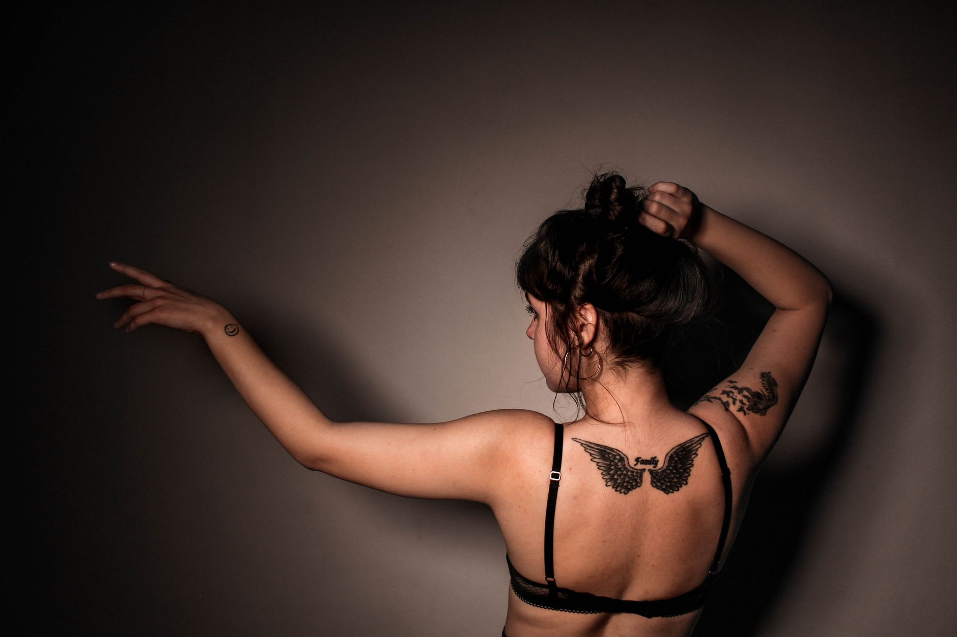 A woman with tattoos poses in lingerie in a moody, dark studio setting, showcasing body art creativity.