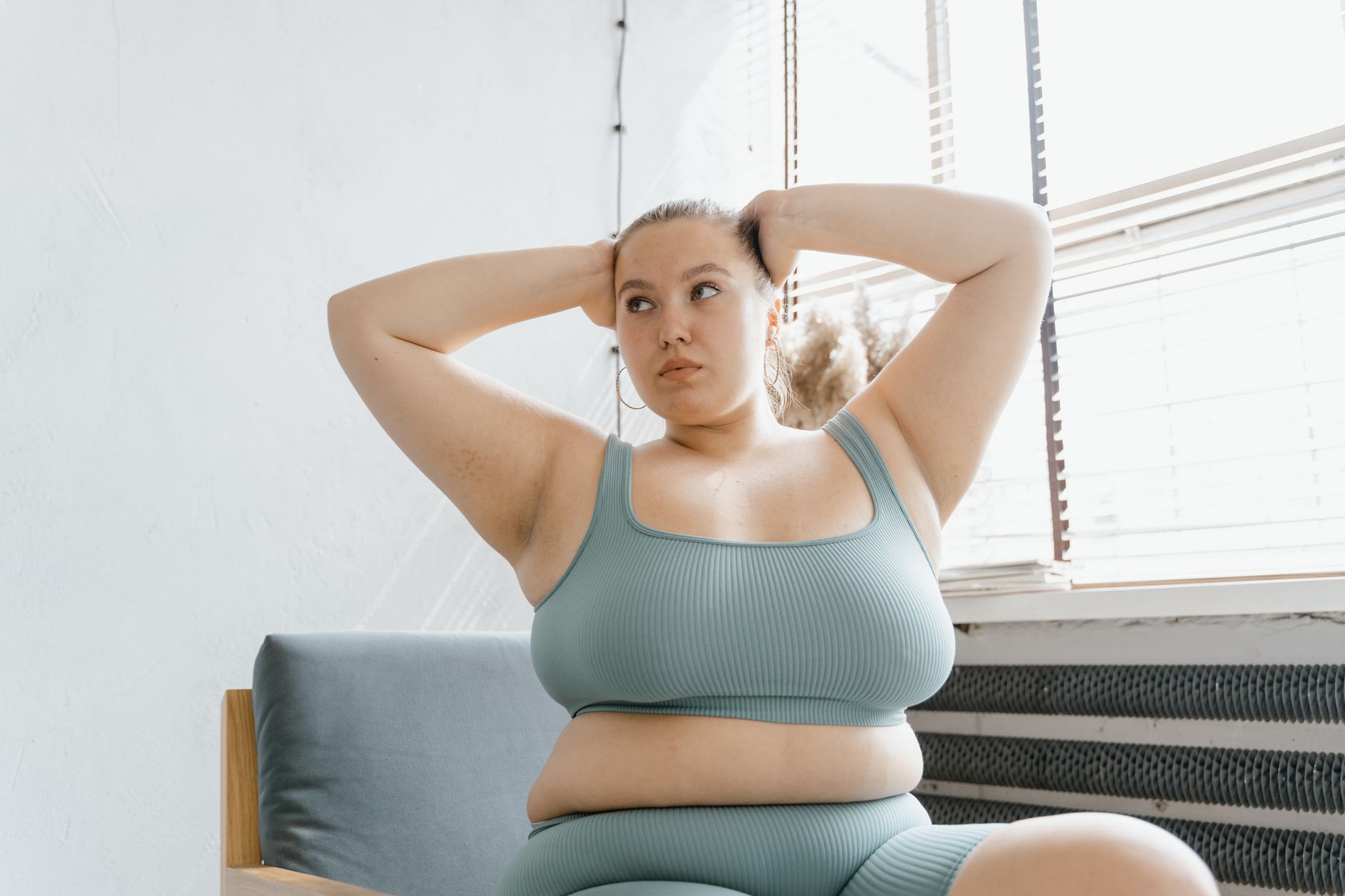 A plus-size woman sitting indoors in a teal outfit, hands on head, looking relaxed by a window.