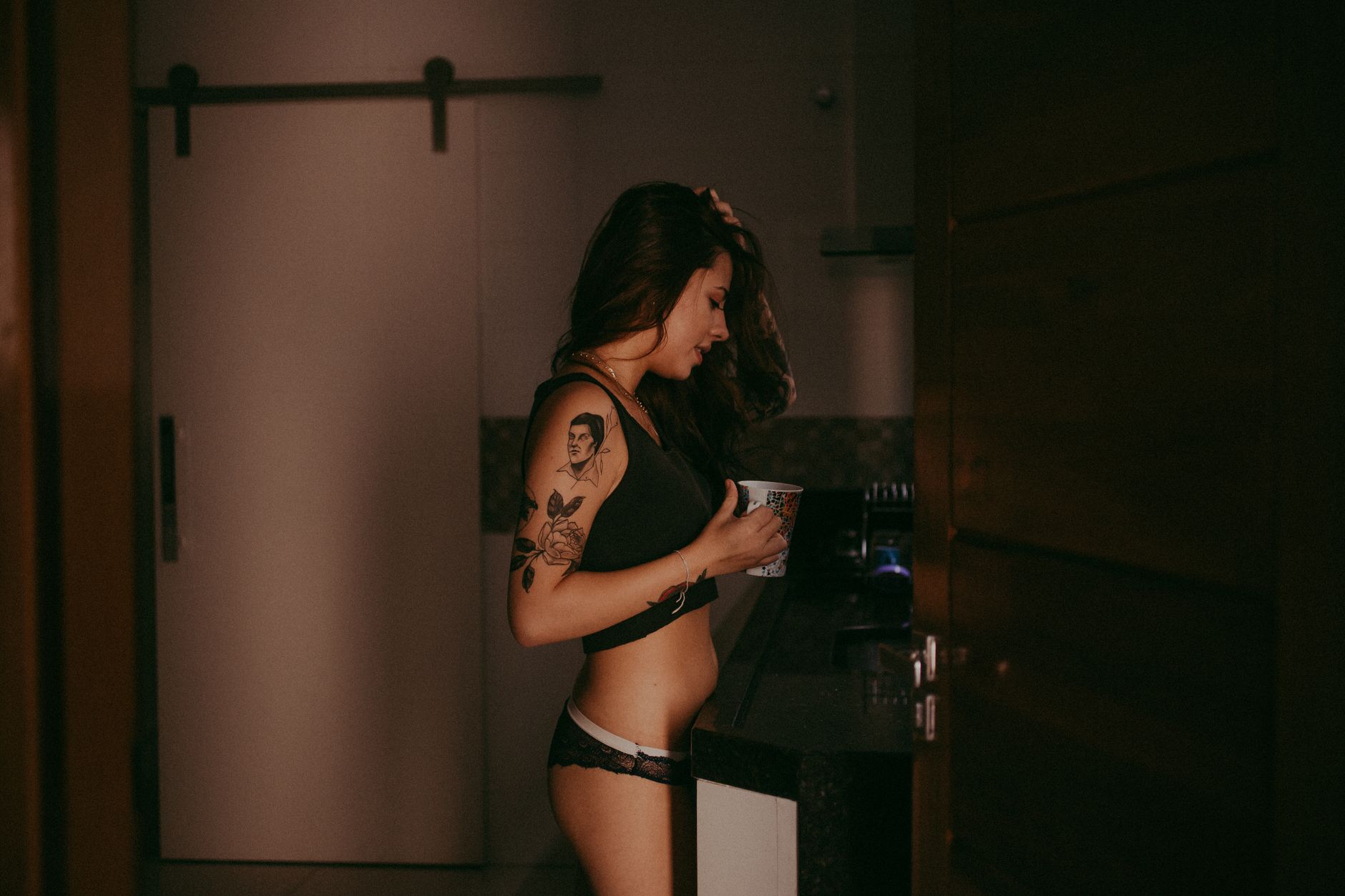 A young woman with tattoos, wearing lingerie, enjoys a casual moment indoors with a cup in hand.