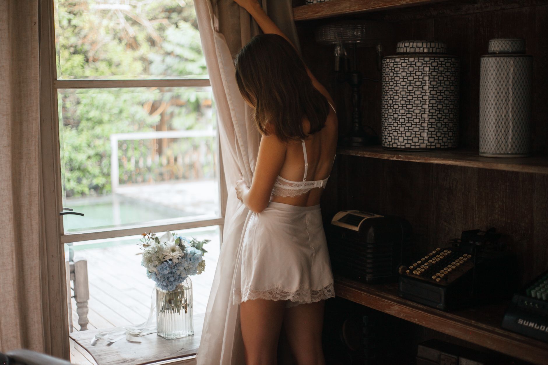 A woman in lingerie gazes outside, surrounded by elegant room decor and flowers.