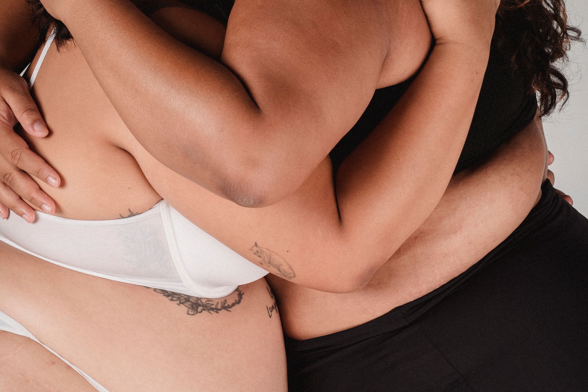 Unrecognizable plump female with tattoos on body wearing bra and panties embracing while standing in light studio on white background