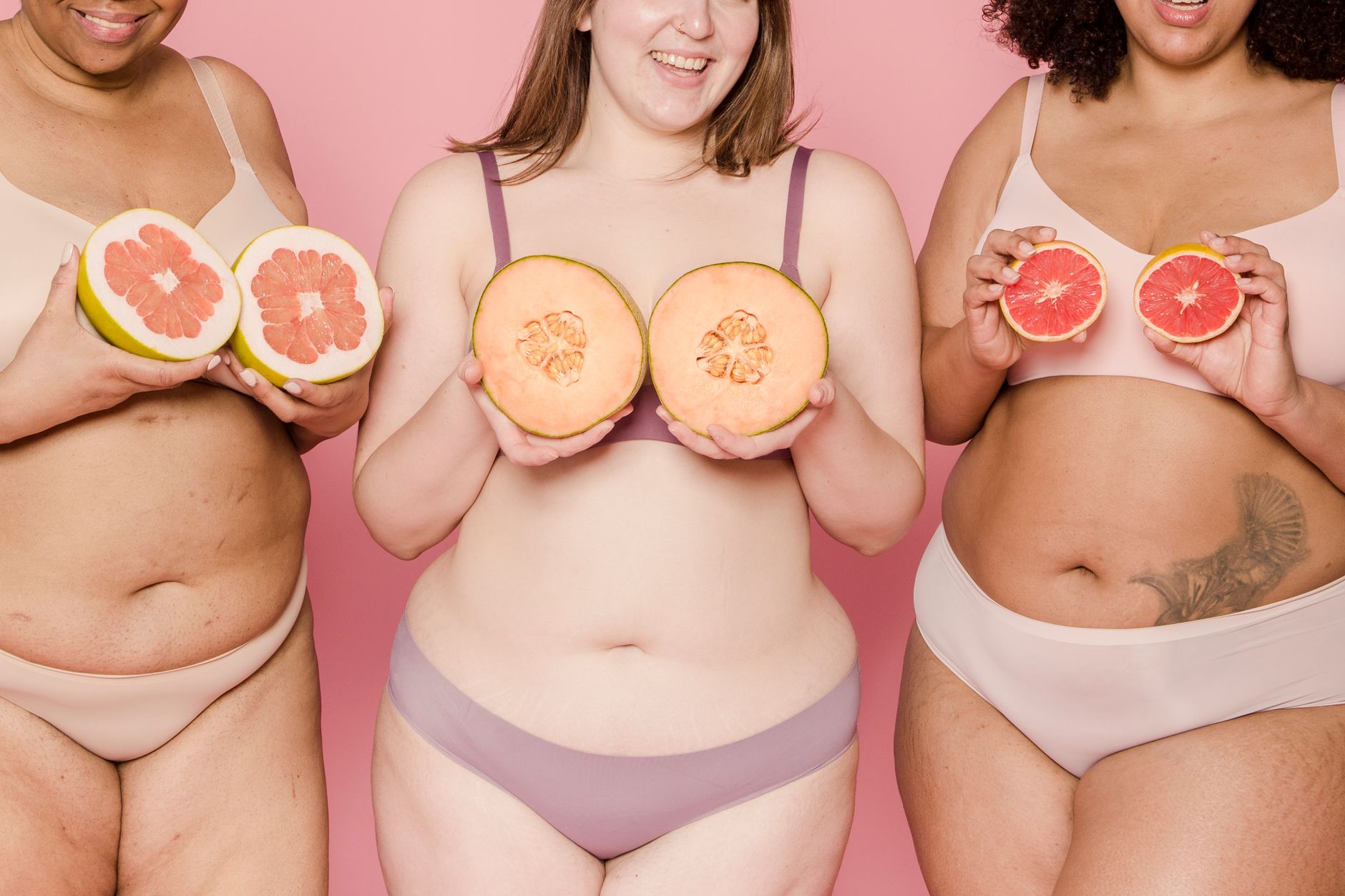Three diverse women in lingerie holding grapefruit and melons on a pink background.