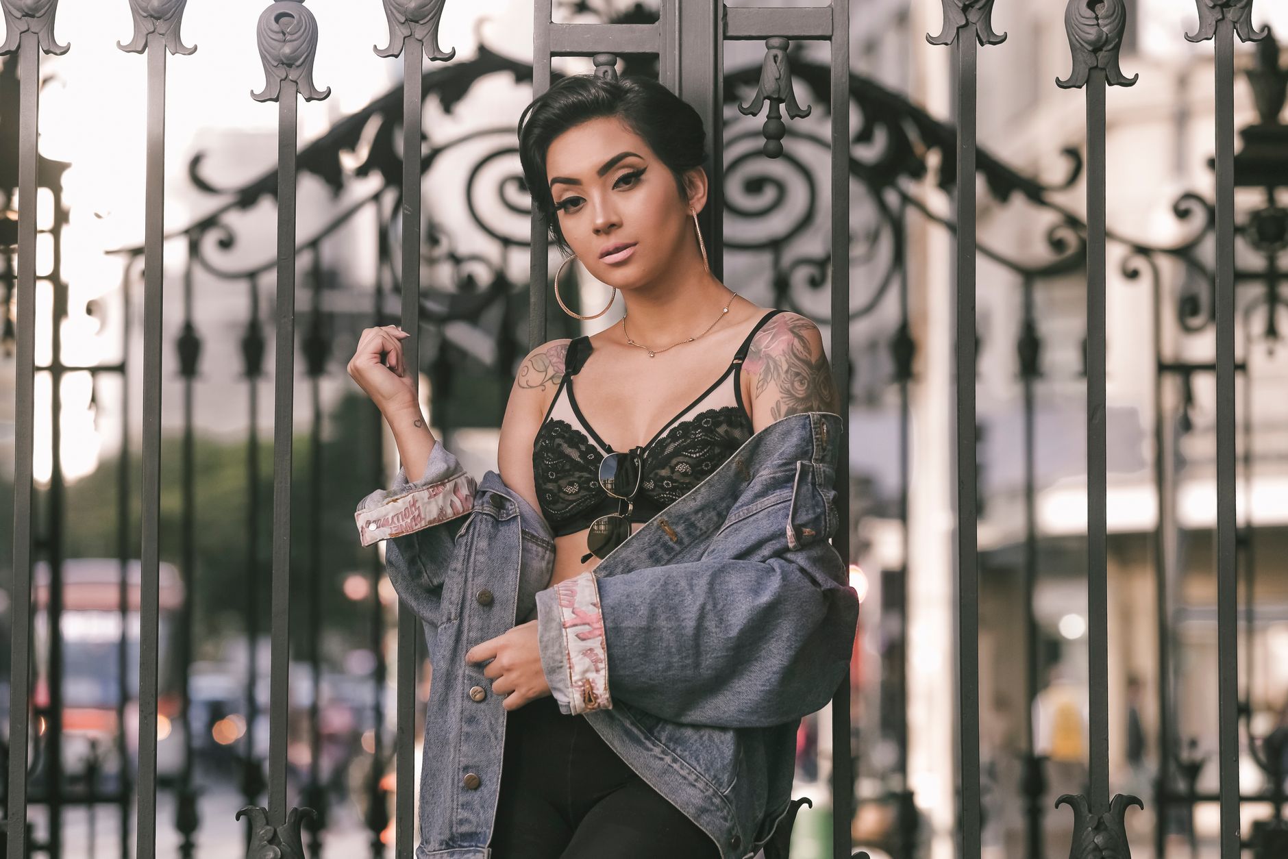 Fashionable young woman in denim jacket posing outdoors by ornate gate.
