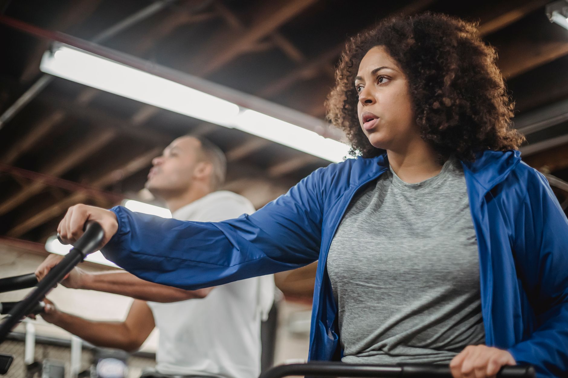 Motivated plus sized African American female in activewear exercising on stationary bike near fit sportsman and looking away while working out in gm