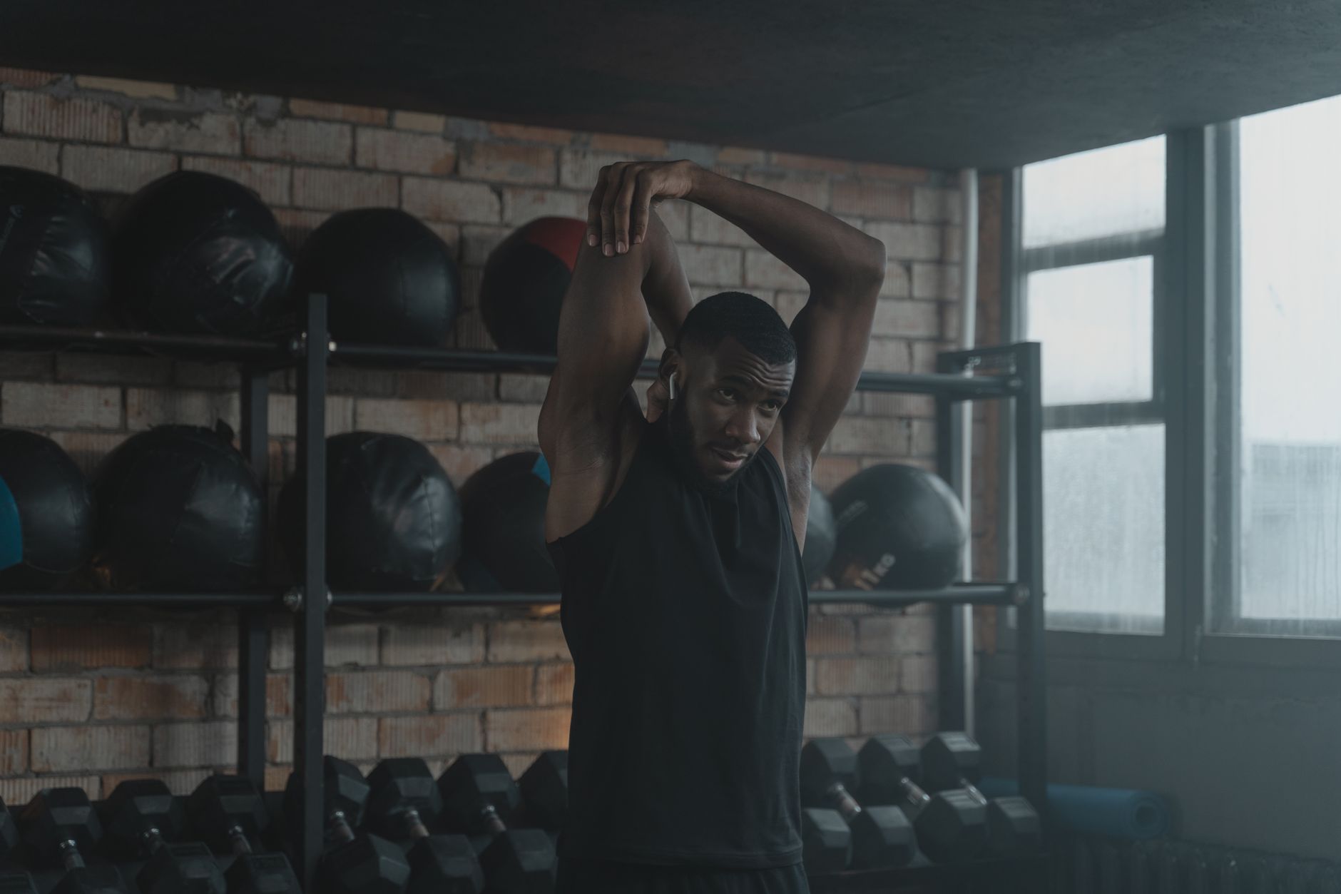 Black man performing an overhead tricep stretch in a gym with exercise equipment in the background.