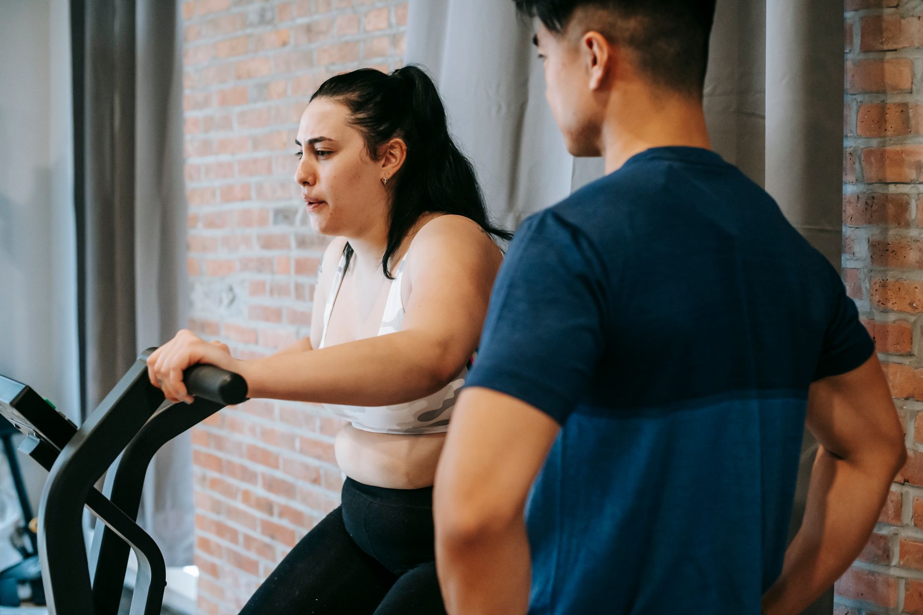 Side view of serious concentrated young obese female doing weight lose exercise on cycling equipment during cardio training with crop ethnic male personal coach