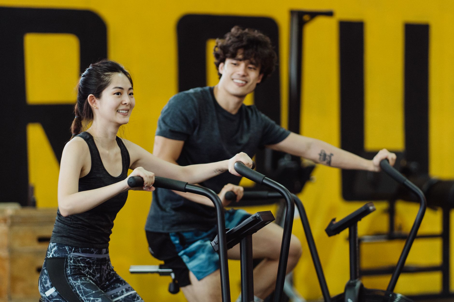 Happy Asian couple on elliptical trainers in gym, smiling and working out together.