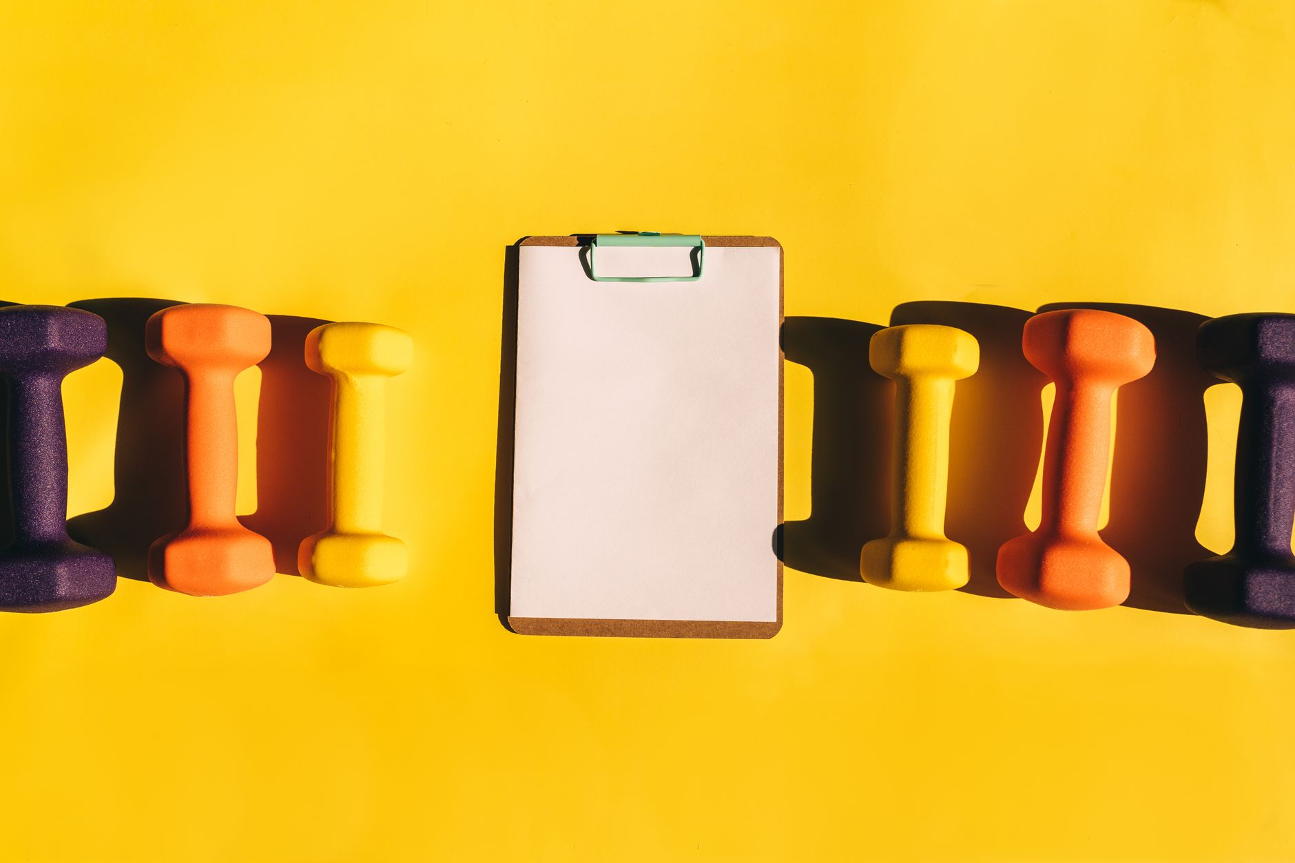 Brightly colored dumbbells arranged with a clipboard on a vibrant yellow background.