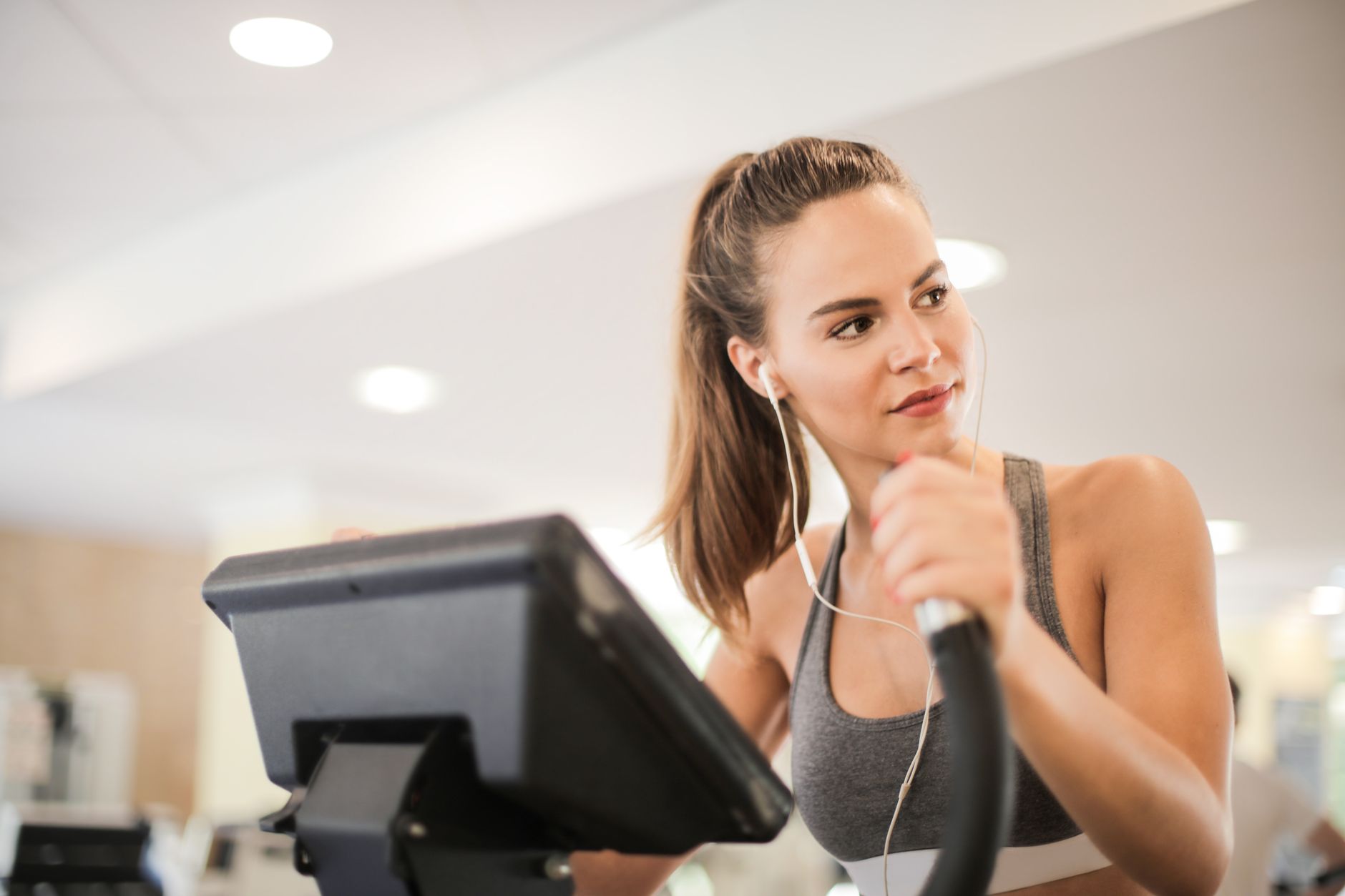 Fit woman working out on an elliptical machine indoors, wearing headphones and a sports bra.