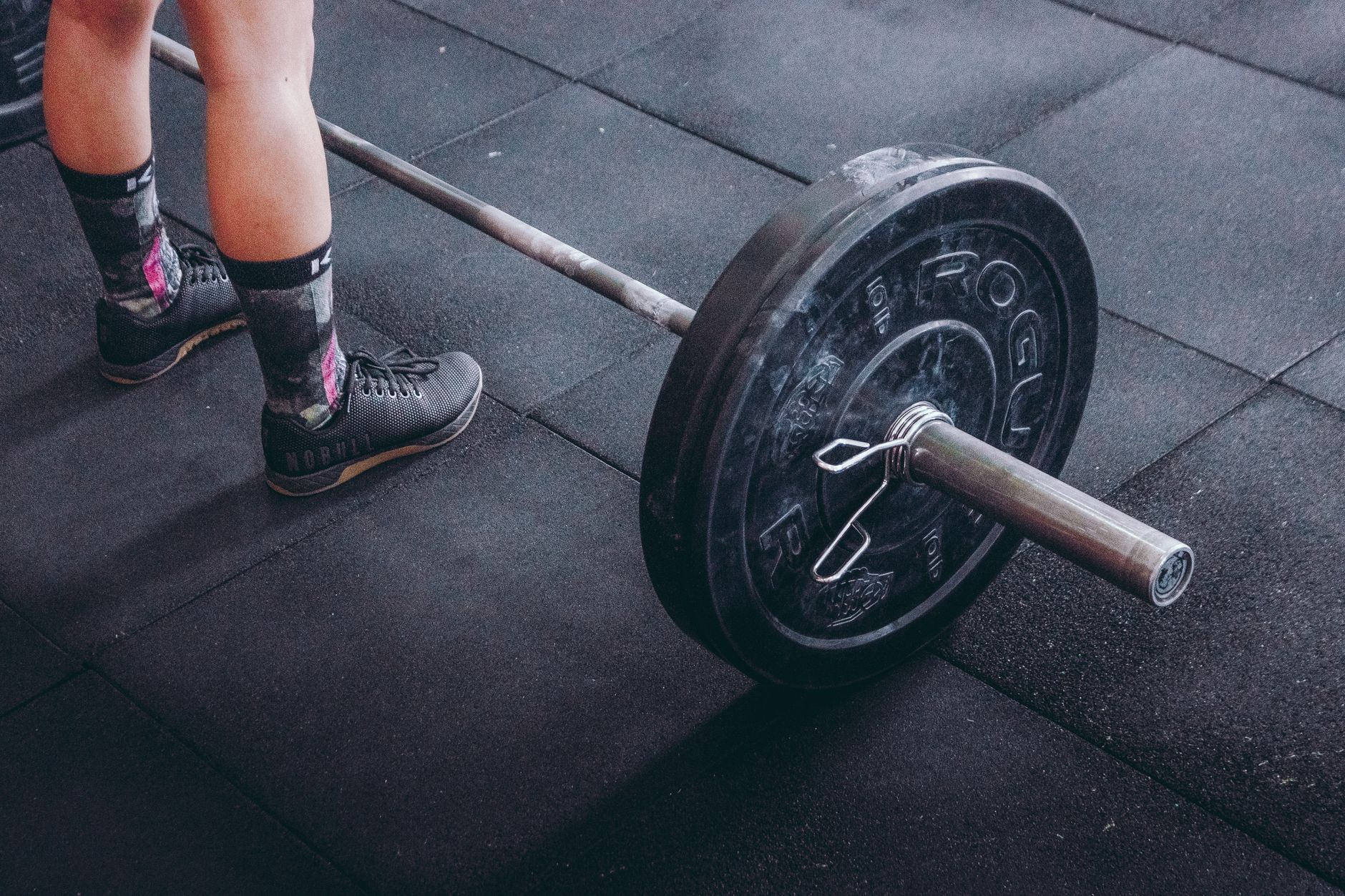 Close-up of an athlete's legs beside a barbell, ready for weightlifting in a gym.