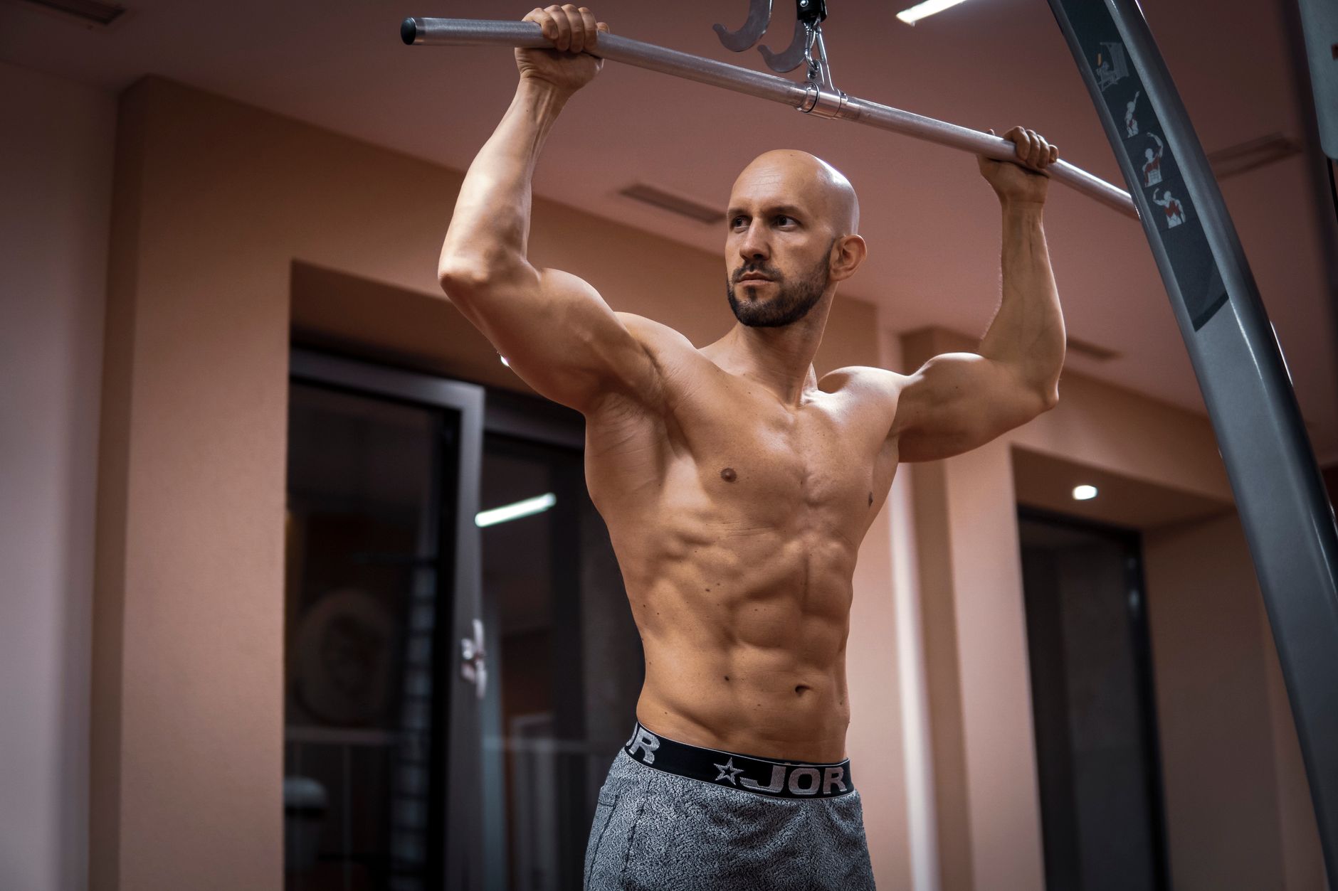 A bald, muscular man performing exercise on gym equipment indoors.