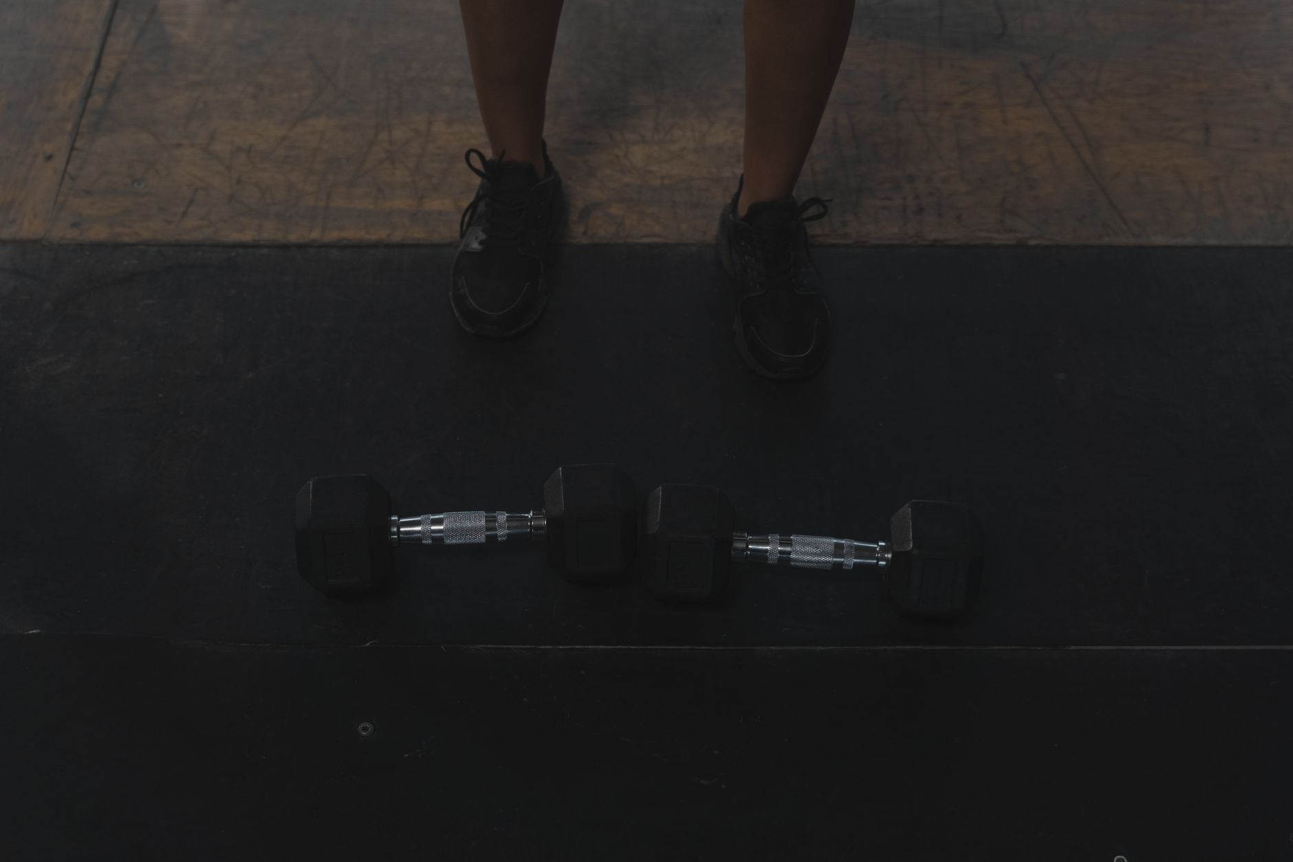 An individual standing on a gym floor behind two dumbbells, ready for a workout.