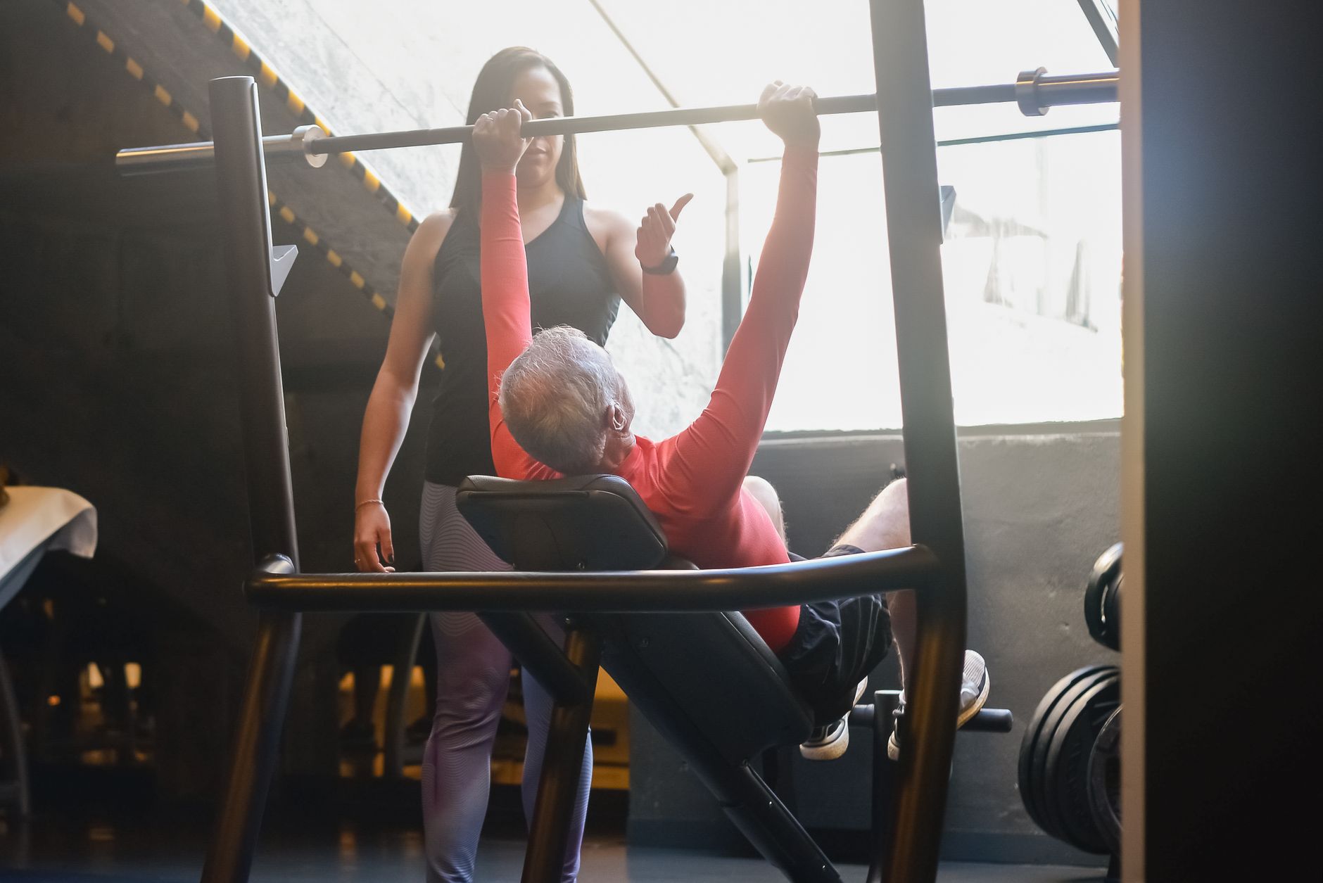 Senior man lifting weights in a gym with guidance from a female trainer, promoting fitness and a healthy lifestyle.