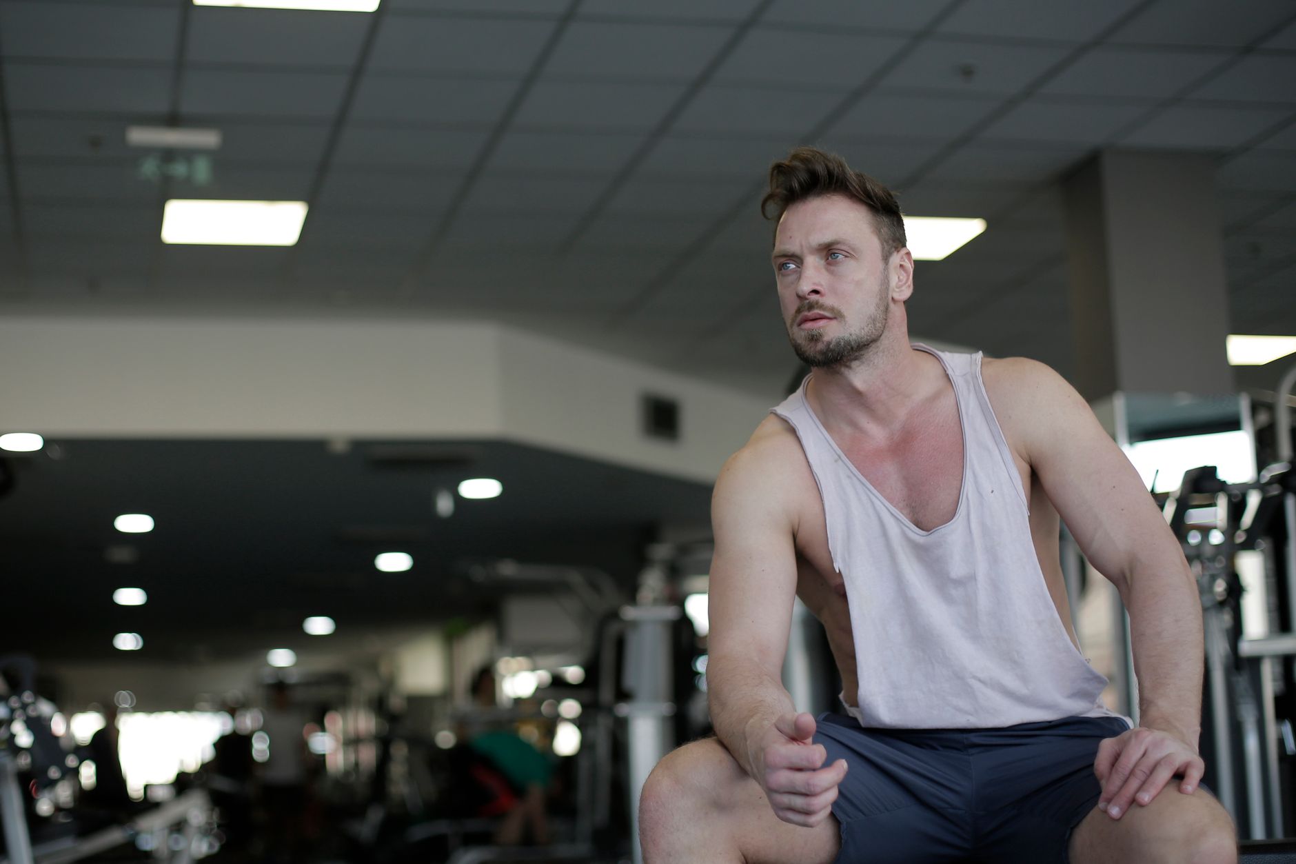 Muscular man with facial hair sitting and looking away in a gym, captured in selective focus.