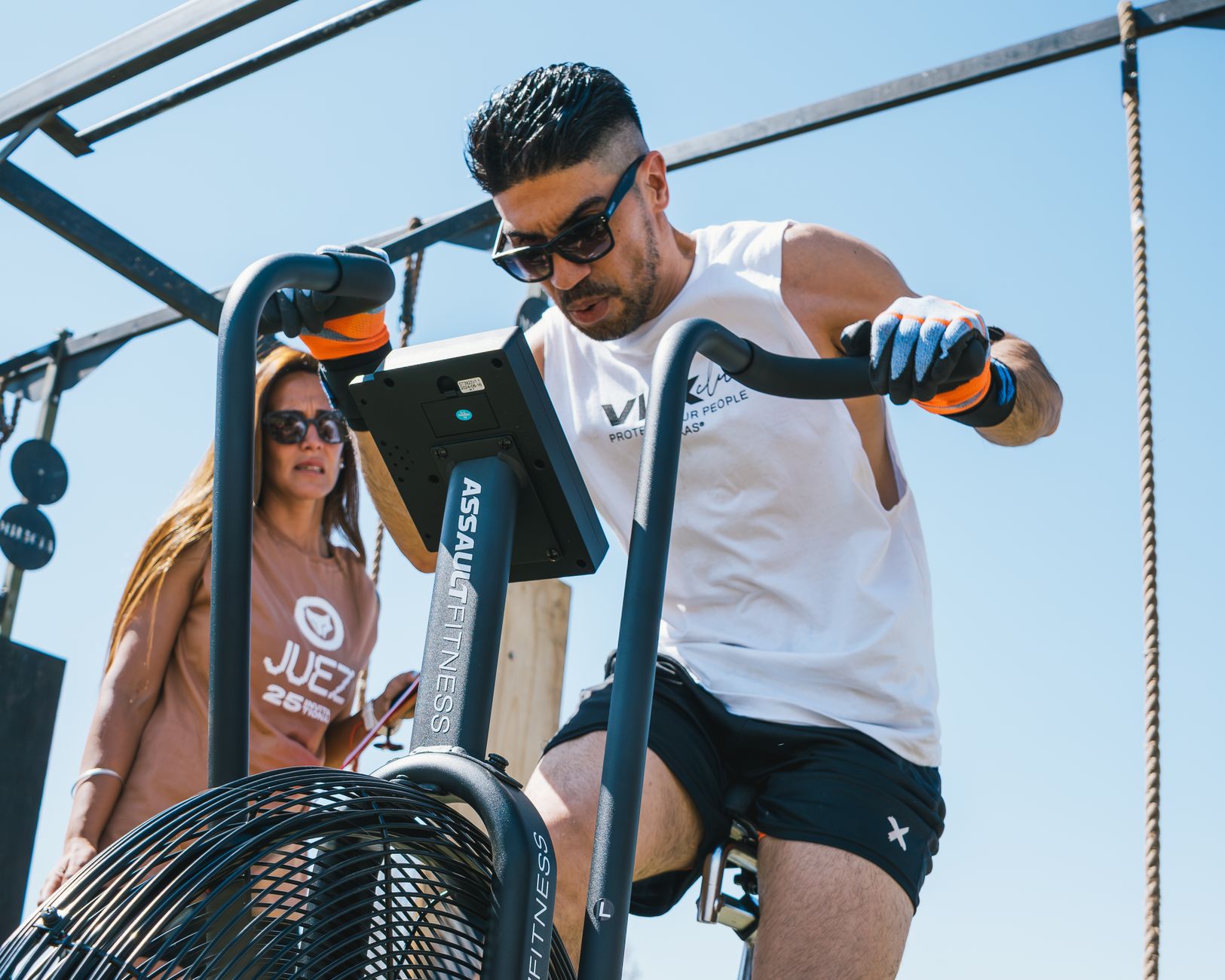 Two adults working out on an air bike outdoors under clear skies.
