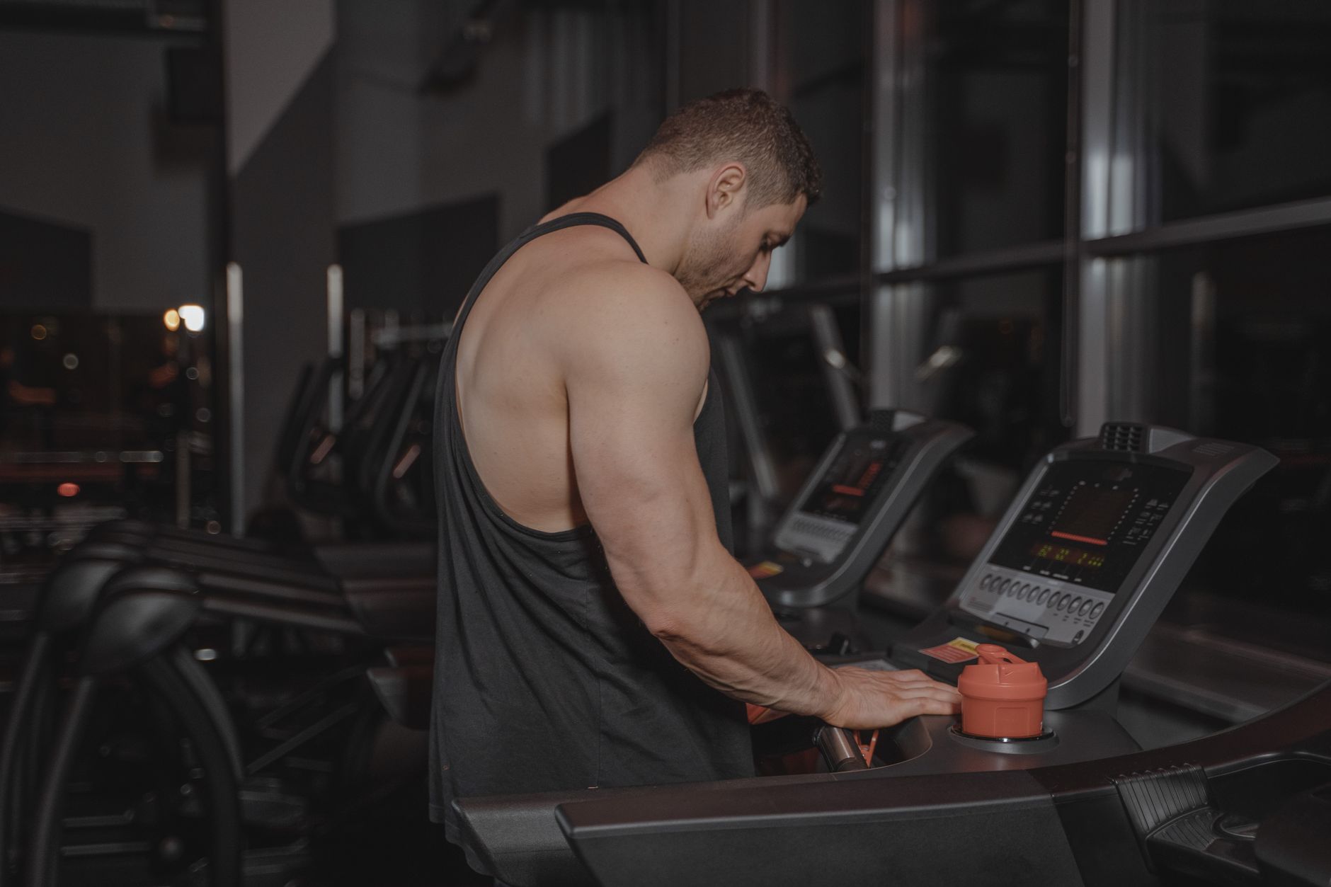 A muscular man adjusting treadmill settings in a modern gym environment at night.