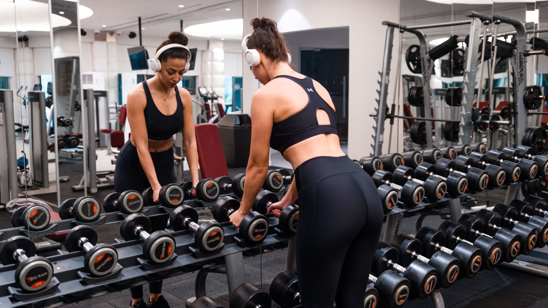 A woman in a gym wearing headphones, holding dumbbells, standing in front of a mirror.