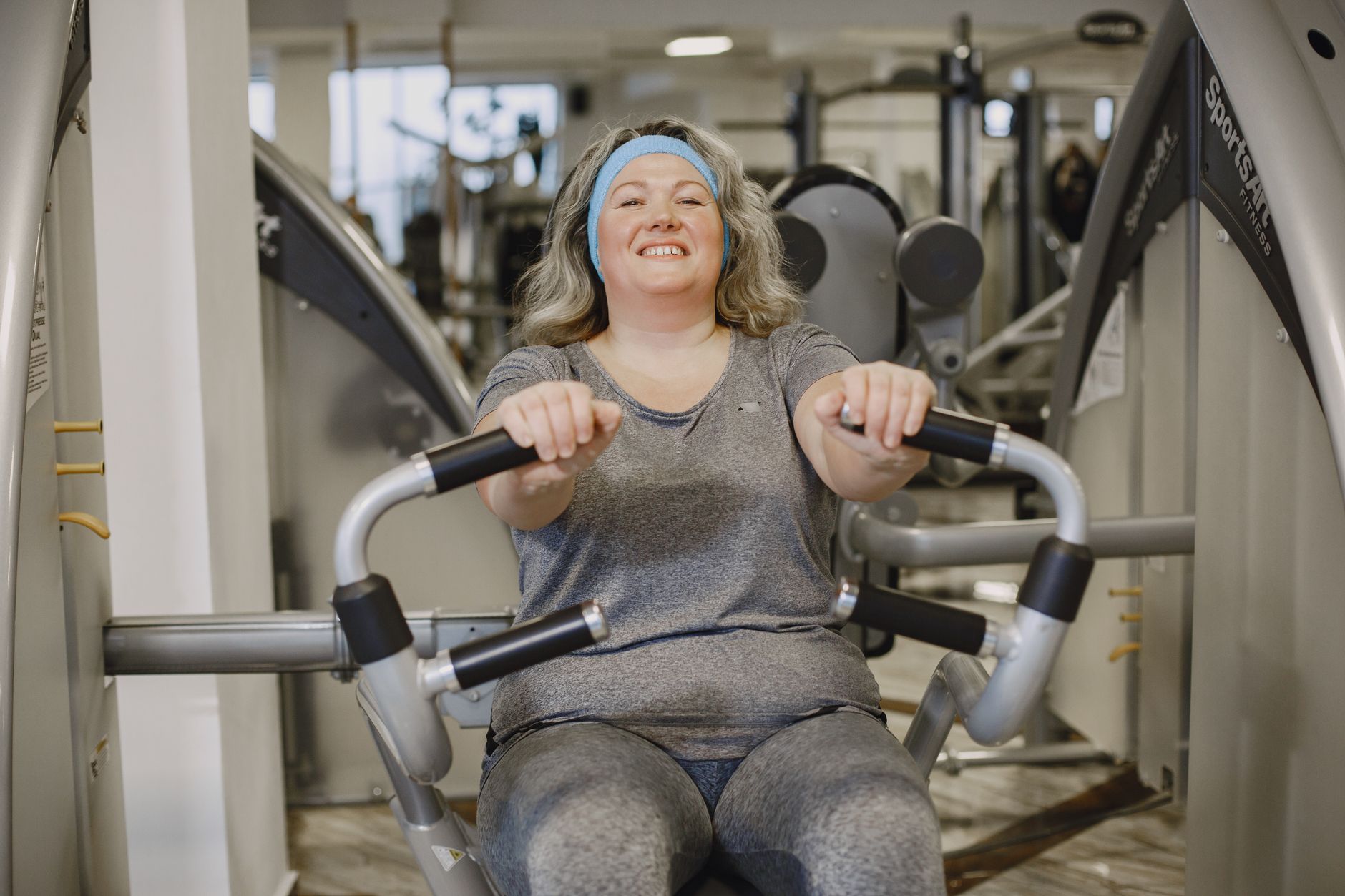 Smiling woman exercises on a gym machine, promoting fitness and well-being.
