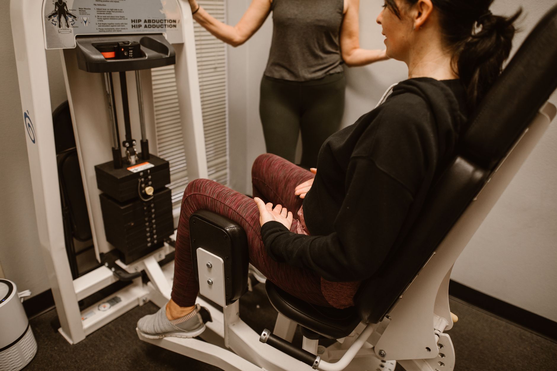 A woman performs exercises on a hip abductor machine with guidance from a trainer.