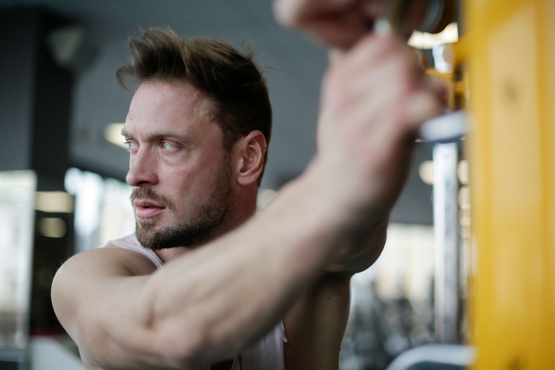 Calm bearded male athlete standing with hands on parallel bar during sport training on blurred background of modern fitness center and looking away