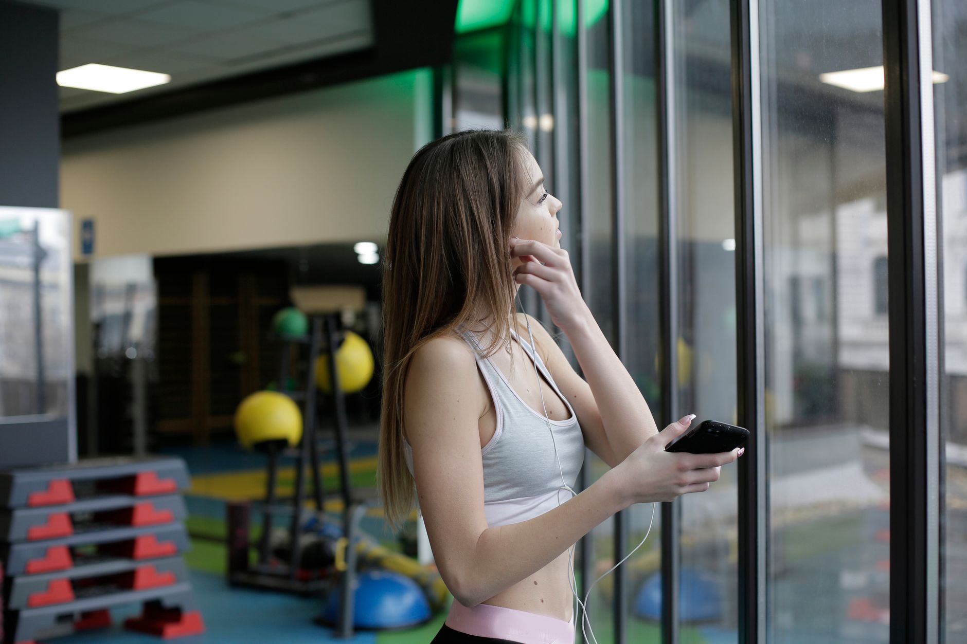 Woman in gym wear looking out window, holding a smartphone and earphones.