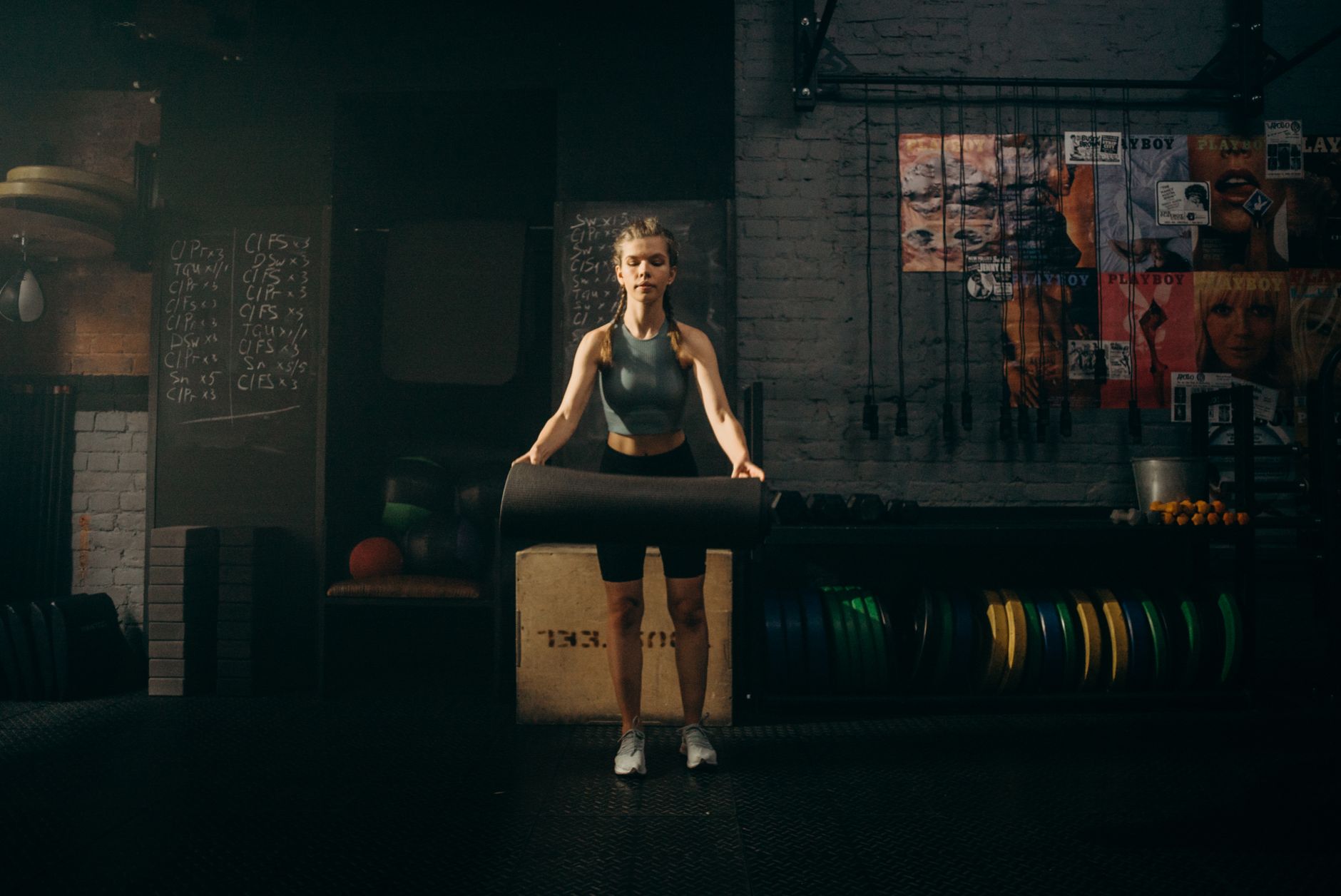 A woman stands in an industrial-style gym, unrolling a yoga mat for her workout.