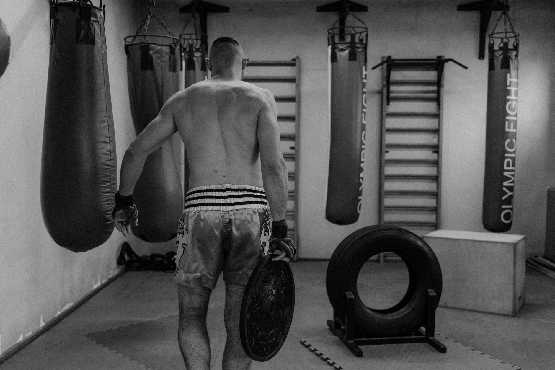 Monochrome image of shirtless man training in a gym with boxing equipment and weights.
