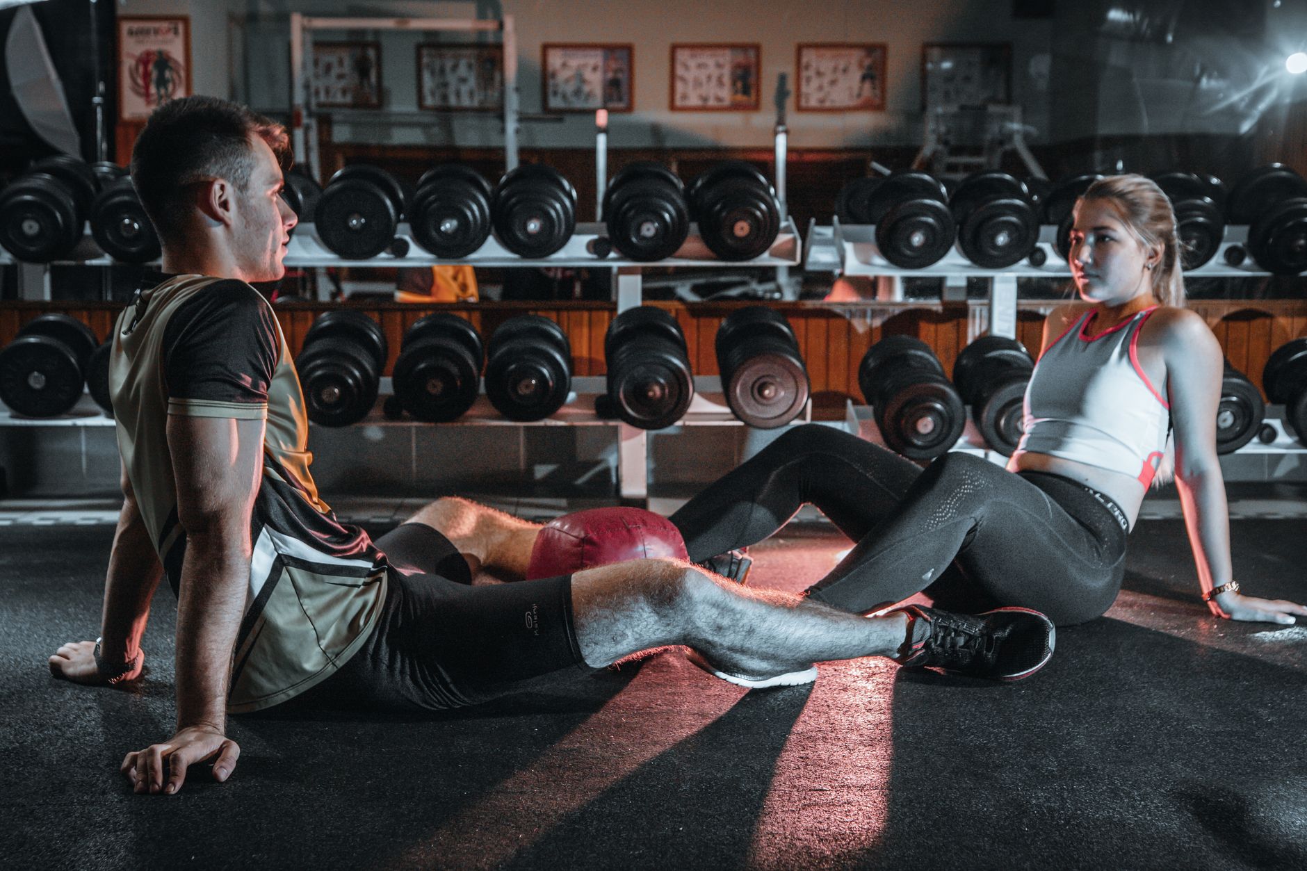 A man and woman resting together on the gym floor, surrounded by dumbbells.