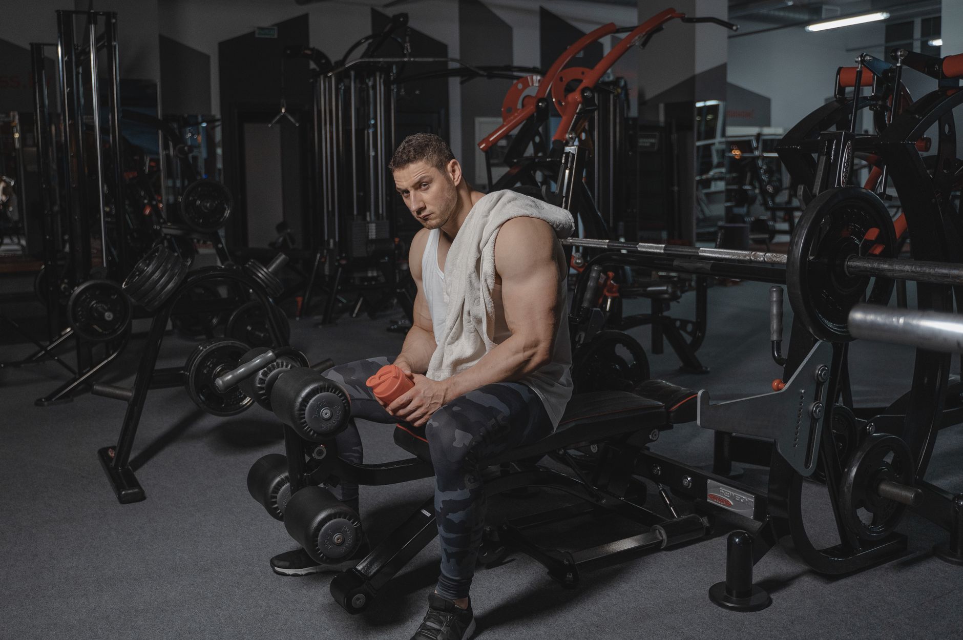 Man seated on bench in gym holding protein shaker, surrounded by equipment.