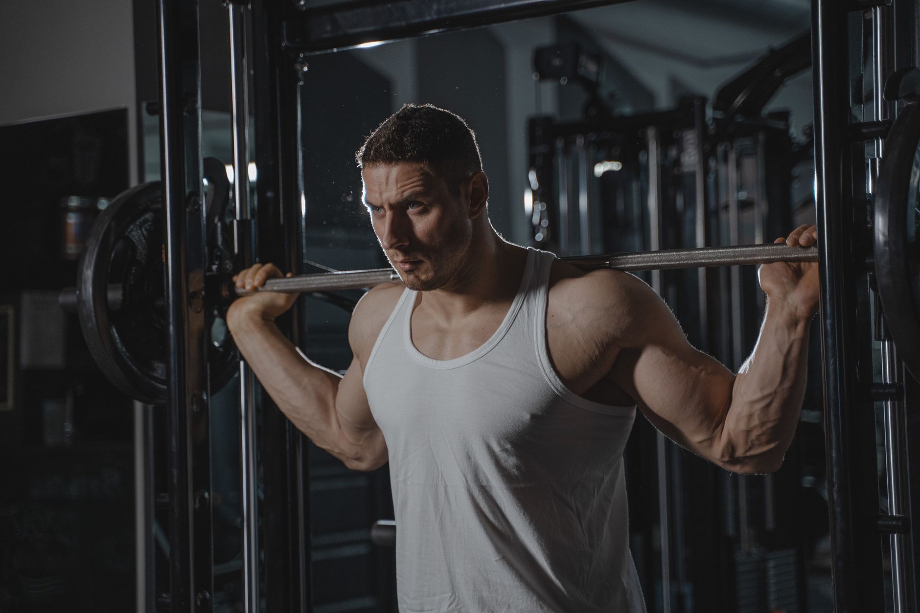 Strong man lifting weights at gym wearing a white tank top, showcasing muscle strength and fitness.