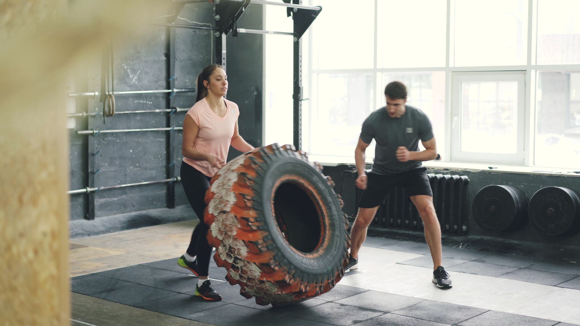 Two adults engaged in a dynamic tire flipping workout inside a gym.