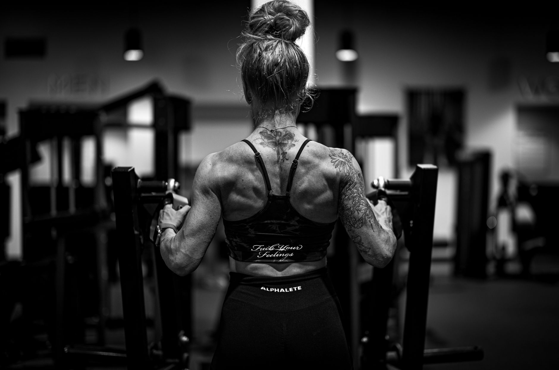 Back view of a tattooed woman lifting weights in a gym, showcasing strength and physique.