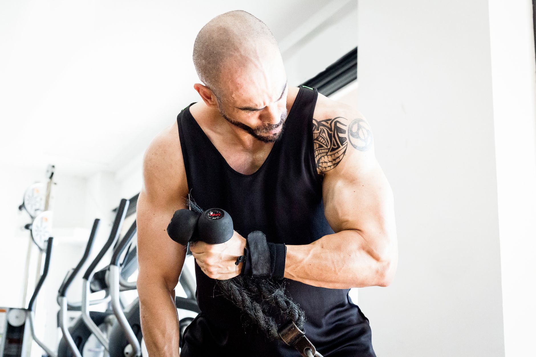 Muscular bald man with tattoos lifting weights in a bright gym.