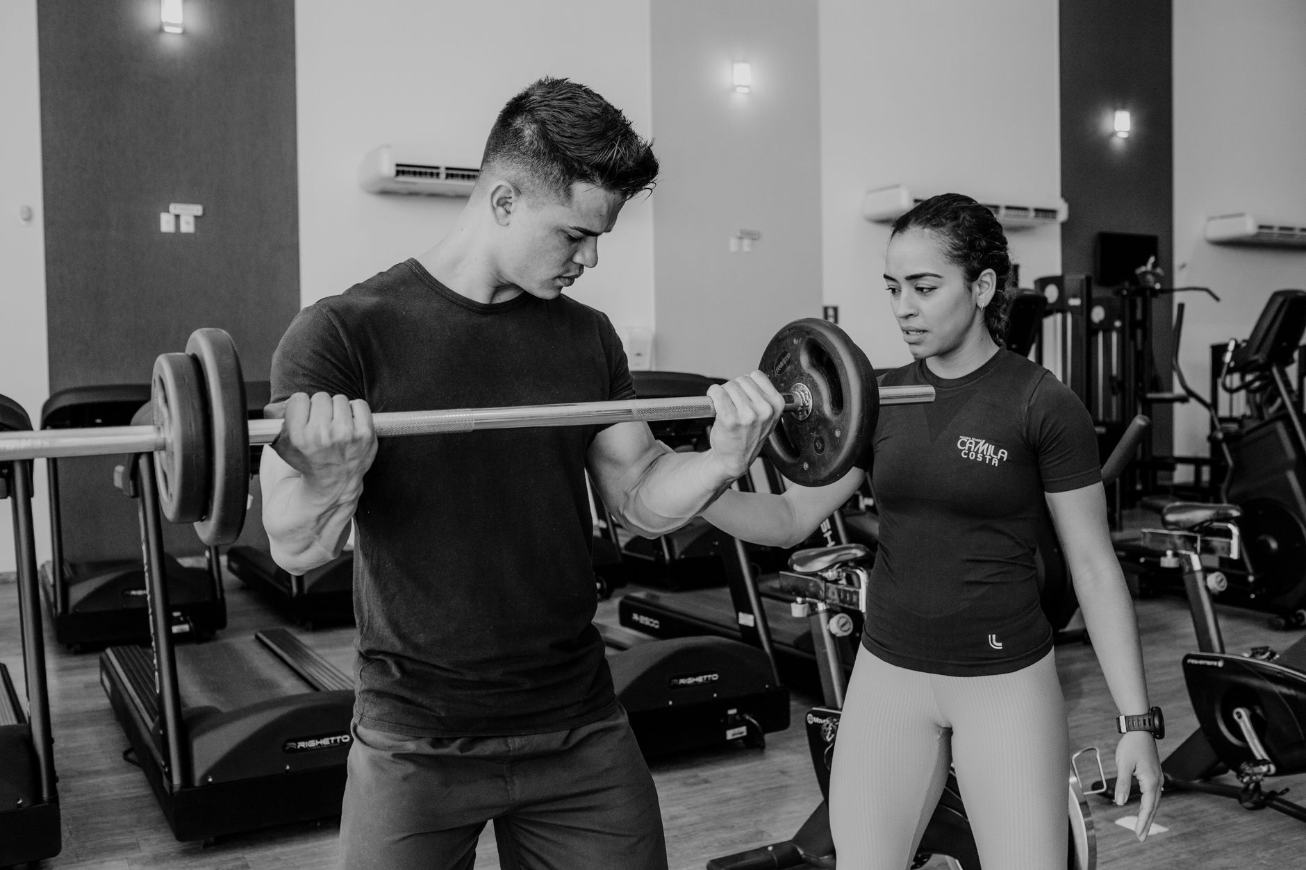 Black and white of strong man training with barbell while trainer supporting in sport club