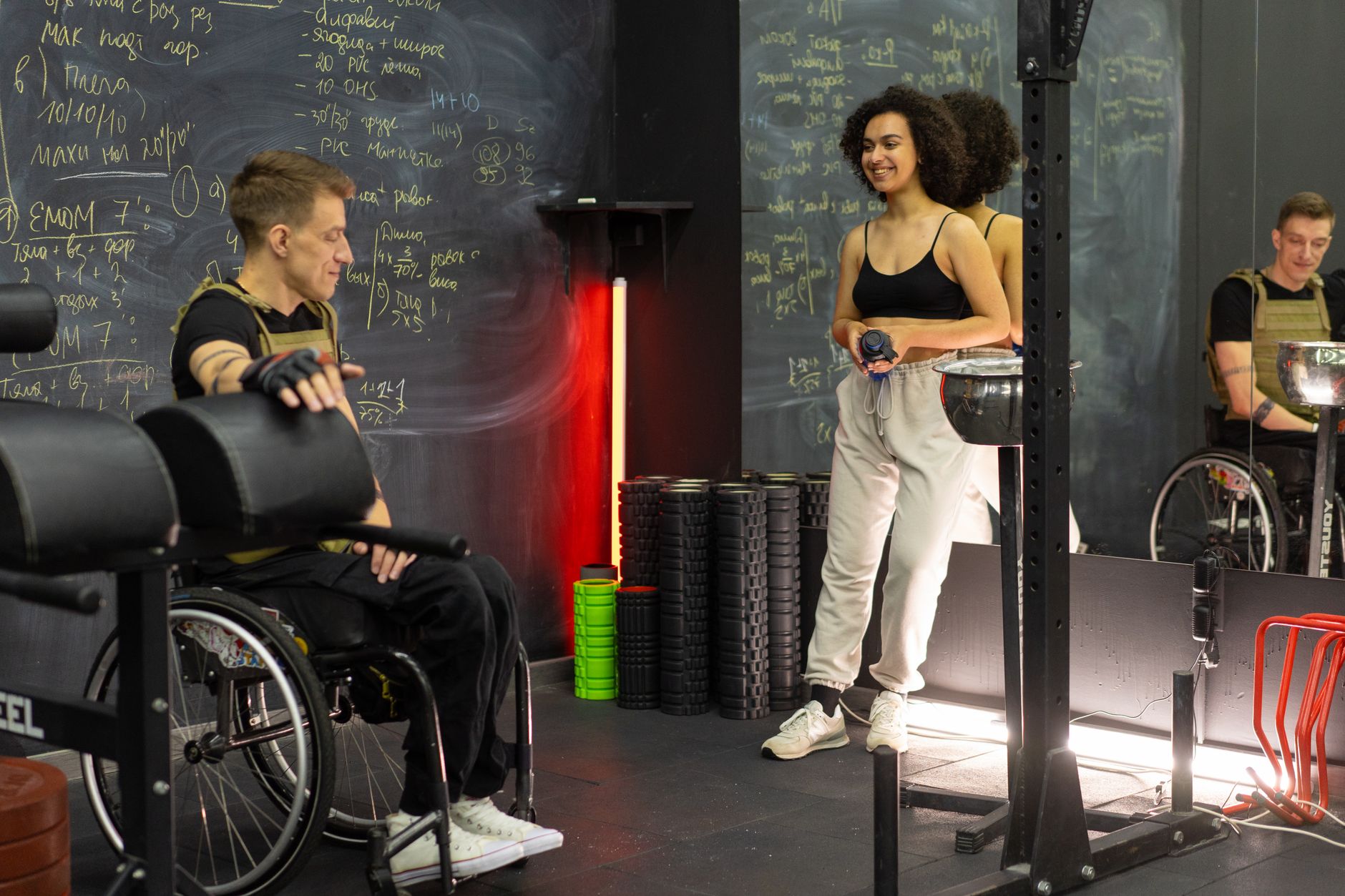 Two people in a gym, one in a wheelchair and one standing, preparing for a workout.