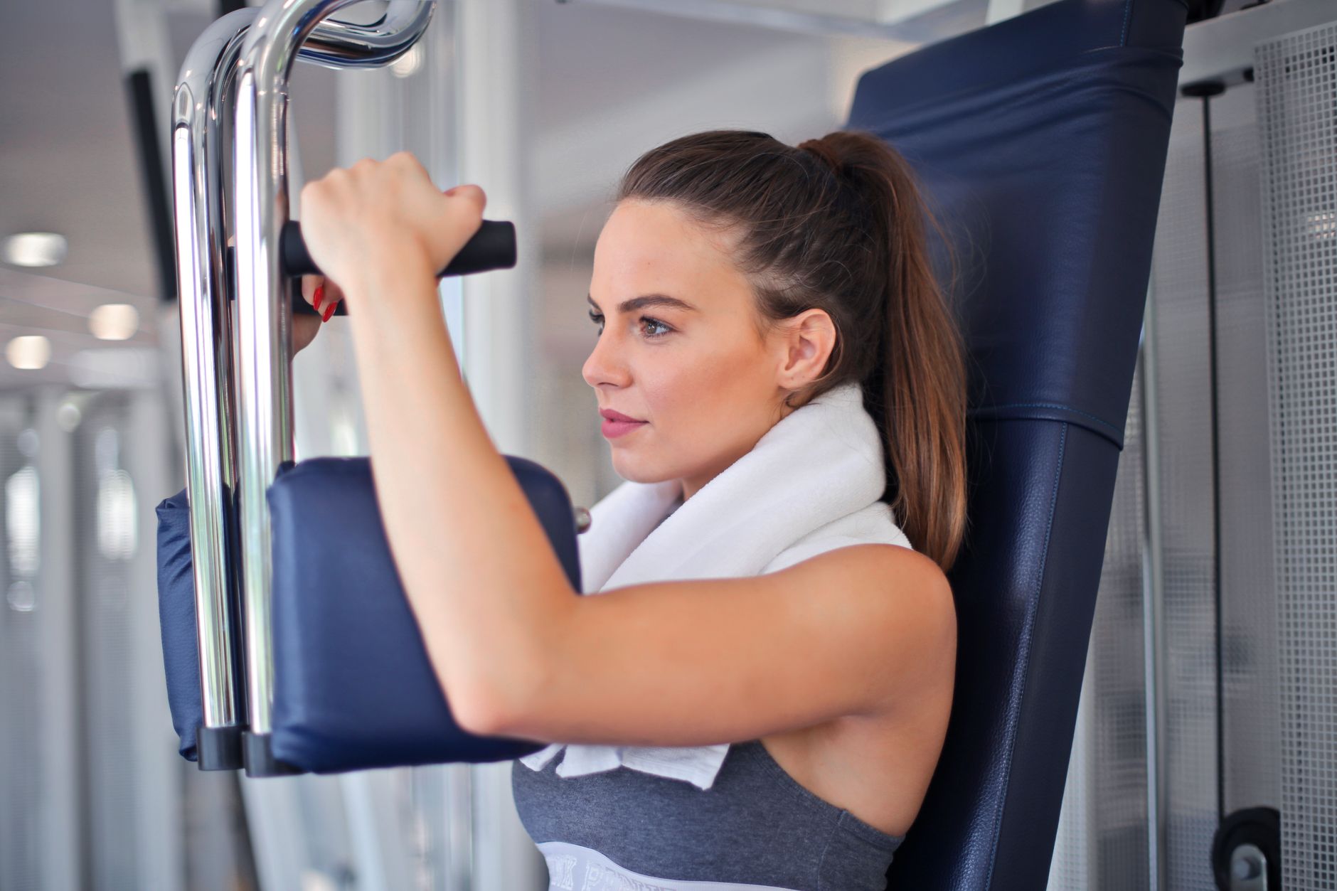 Slender focused woman in sportswear with towel on neck performing exercise on weight machine in light contemporary gym