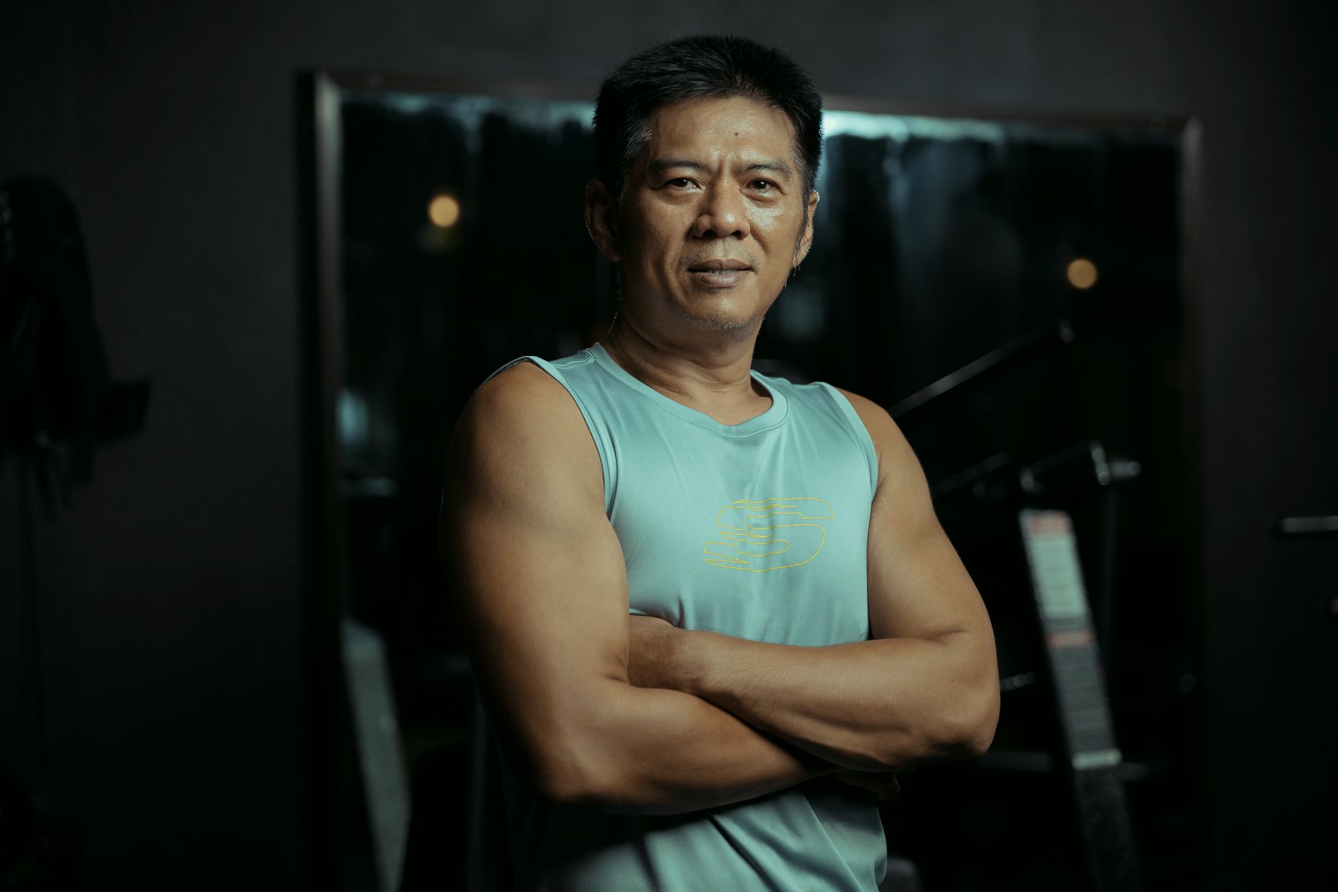 A mature man stands confidently with crossed arms in a gym environment, showcasing fitness and strength.