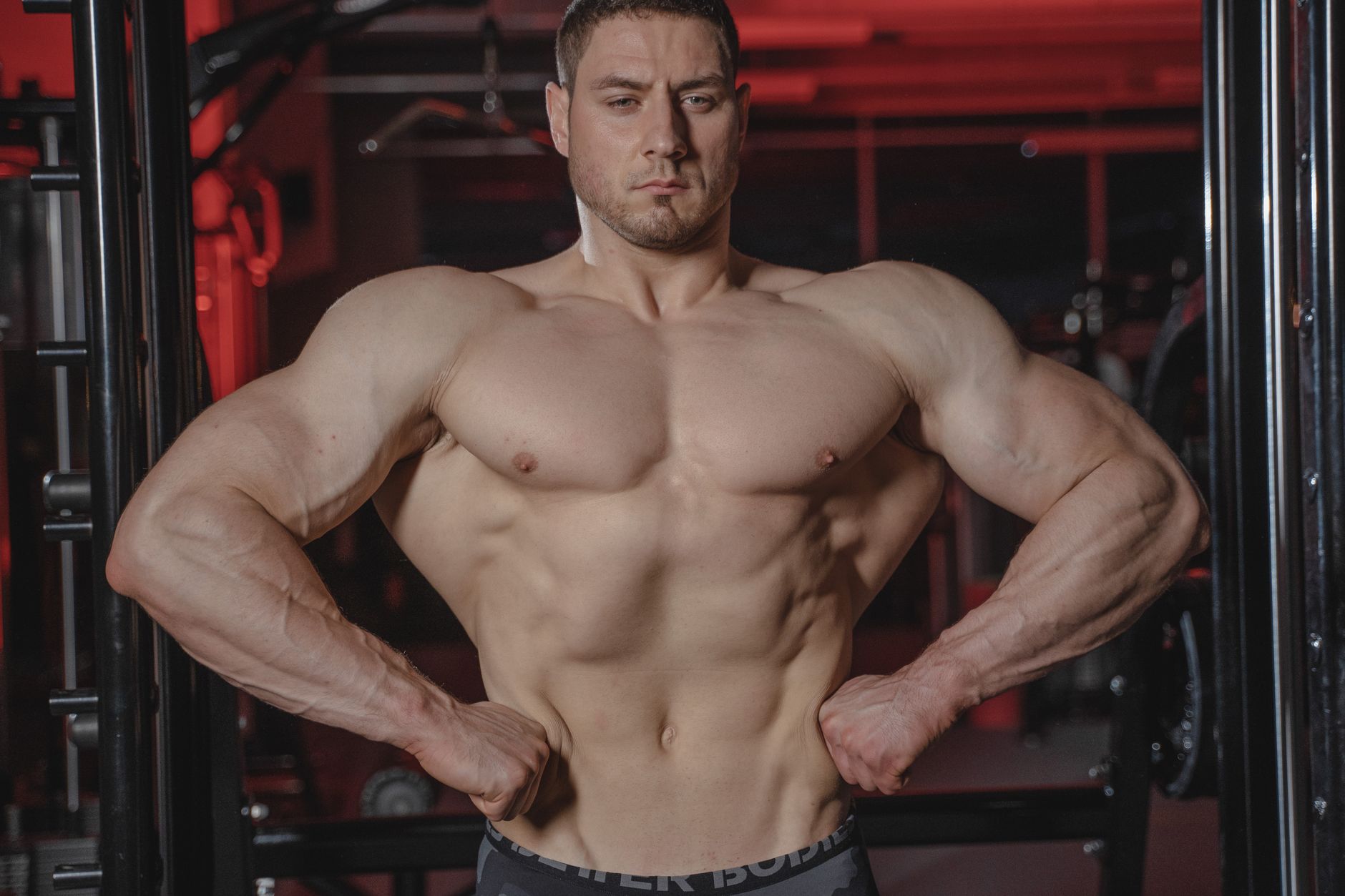 Intense shot of a muscular man posing topless in a gym setting showcasing physique.