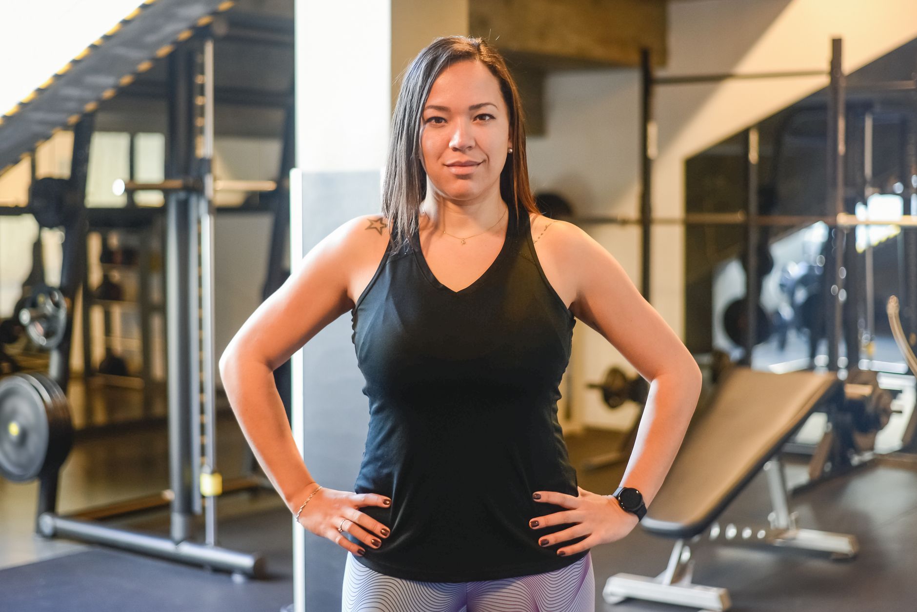 Woman confidently standing in a gym with a blurred background and black tank top.