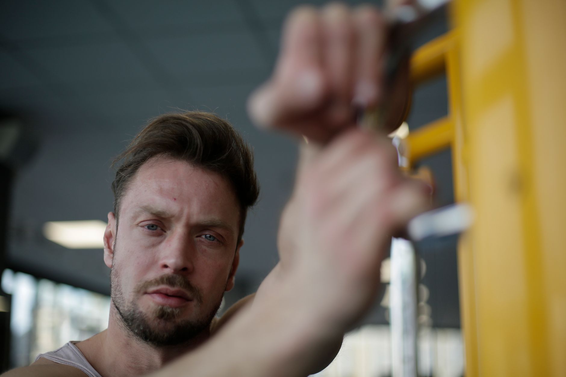 Determined bearded male athlete standing with hands on parallel bar during sport training on blurred background of modern fitness center and looking at camera