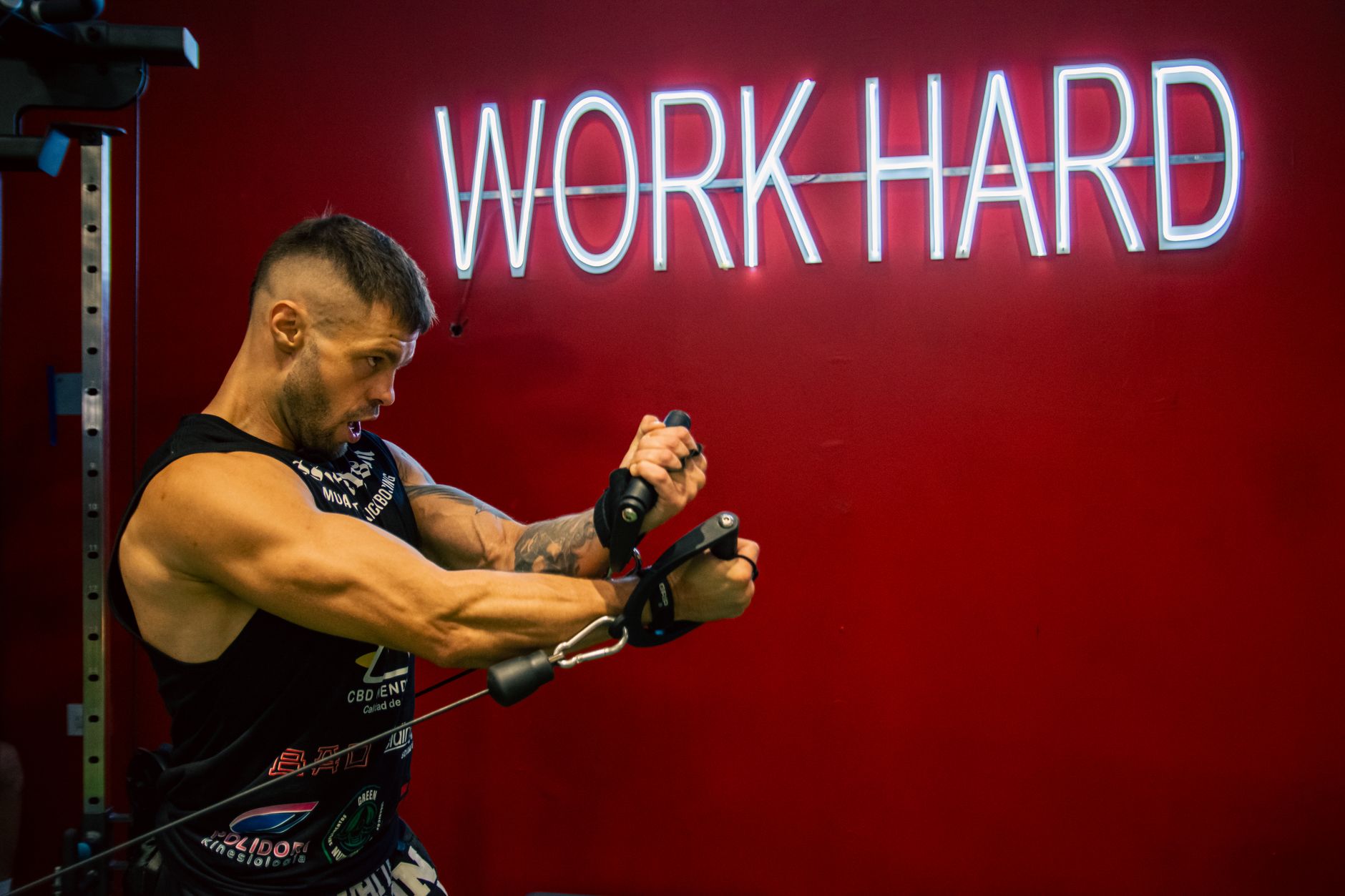 A muscular man exercising in the gym with a motivational 'Work Hard' neon sign on a red background.